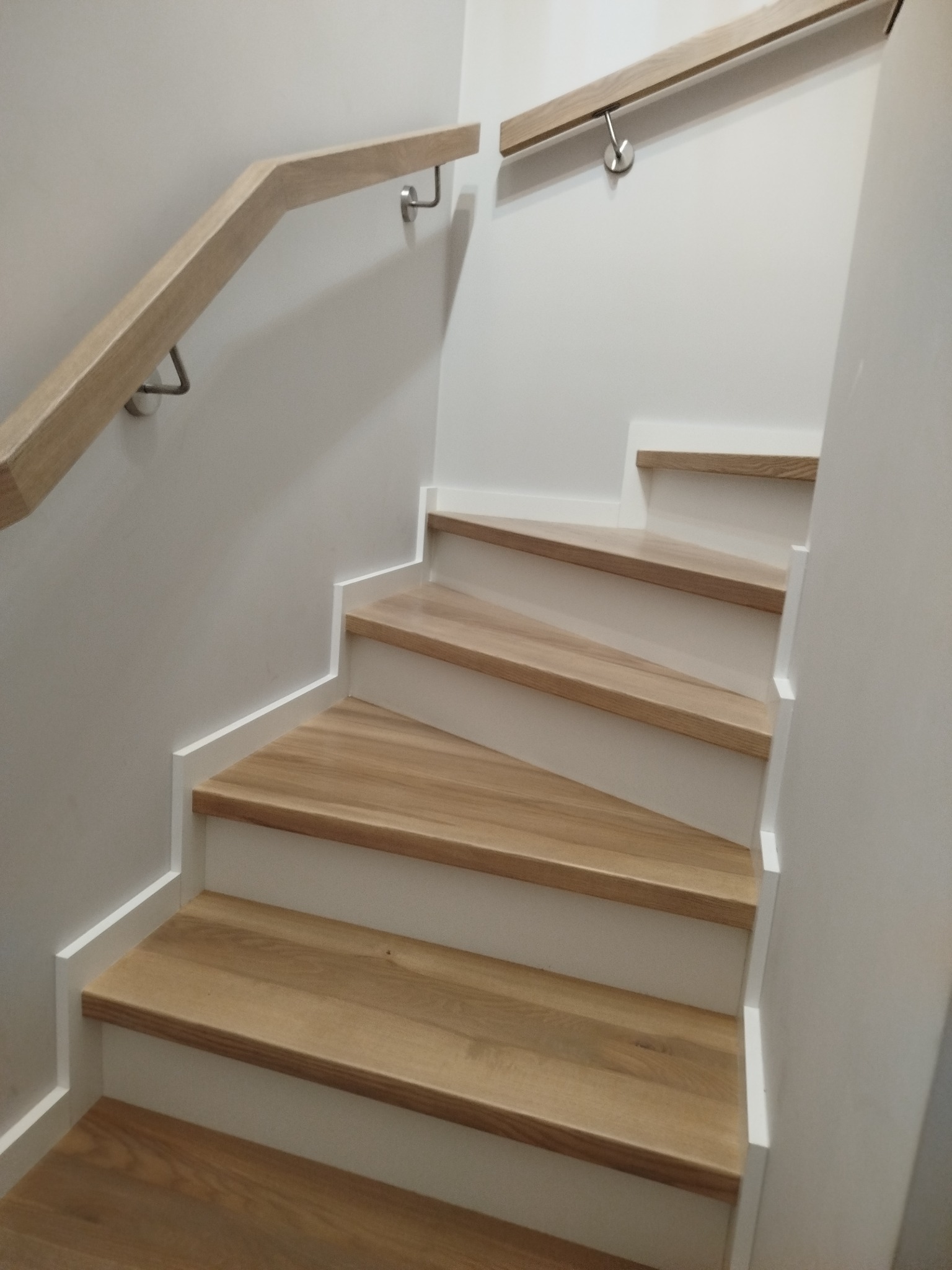 Jasne, drewniane stopnie z białymi podstopnicami i drewnianą poręczą na ścianie. Minimalistyczny design, widok z dołu, oświetlenie naturalne.