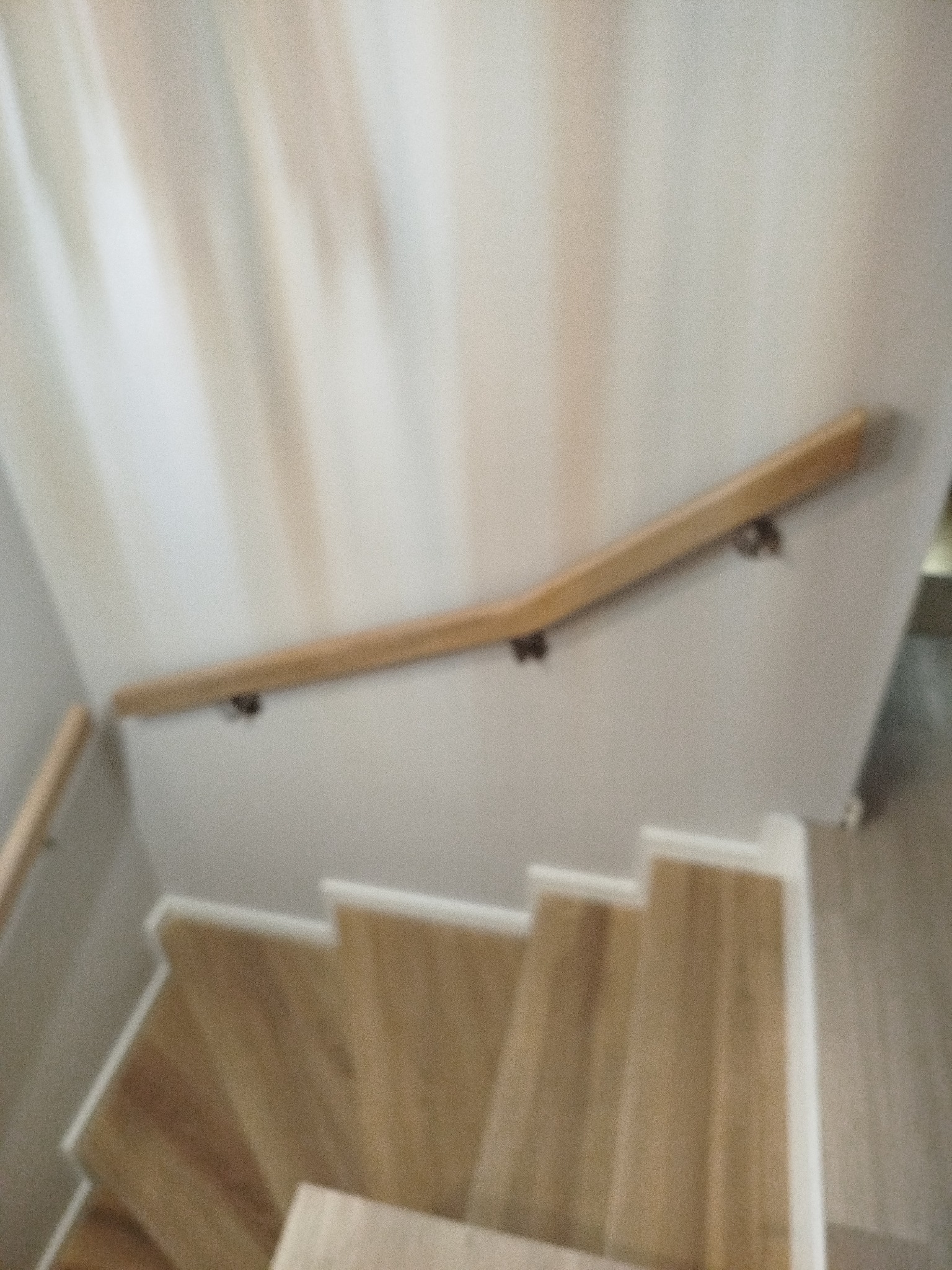 Jasne, drewniane schody z poręczą zamocowaną do ściany. Ujęcie z góry, widoczne stopnie i fragment ściany. Rozmycie dodaje dynamiki.