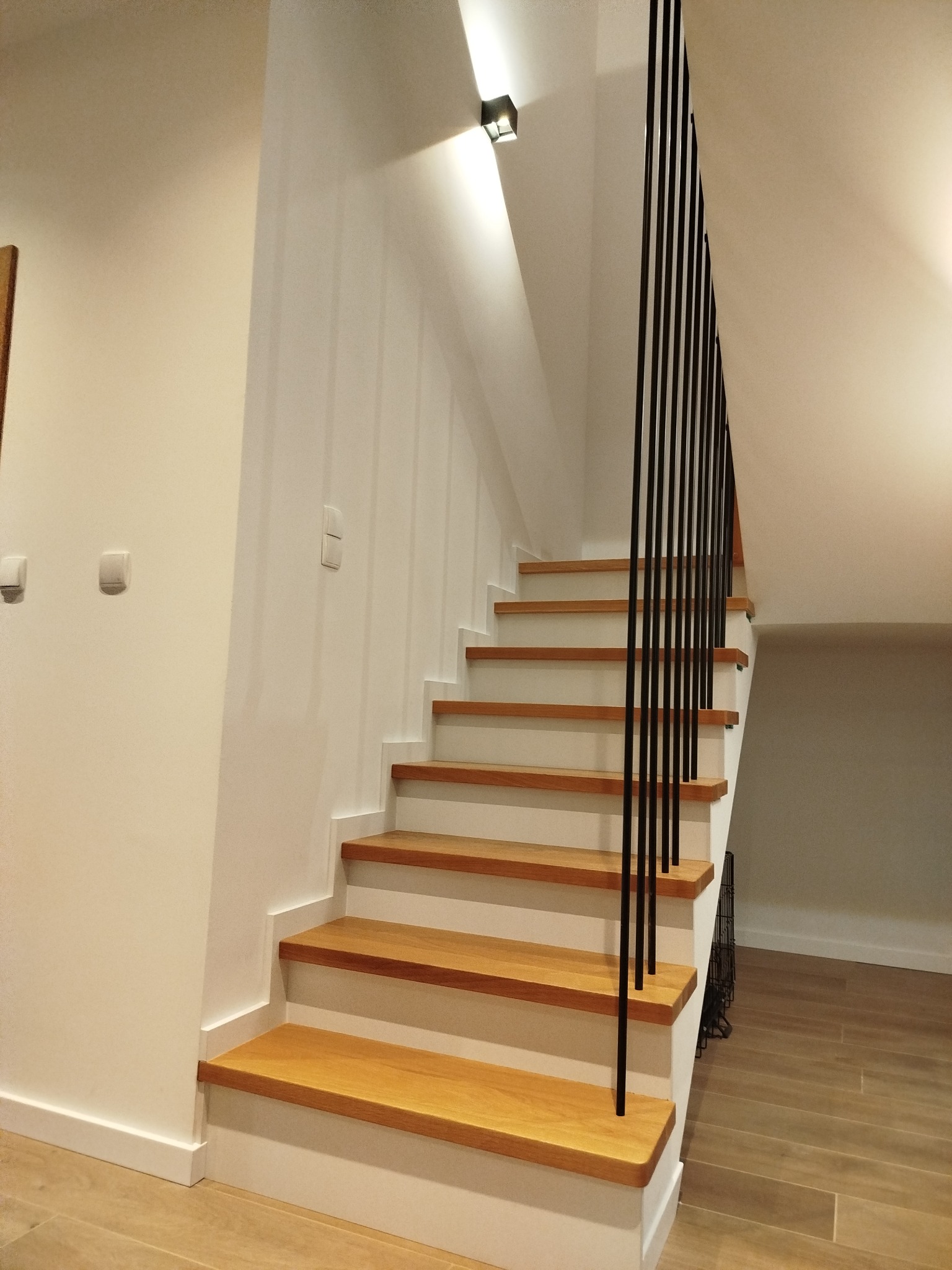 Jasne, nowoczesne schody z drewnianymi stopniami i czarną, minimalistyczną balustradą. Ściany białe, oświetlenie punktowe. Widok z dołu, podkreślający geometrię.