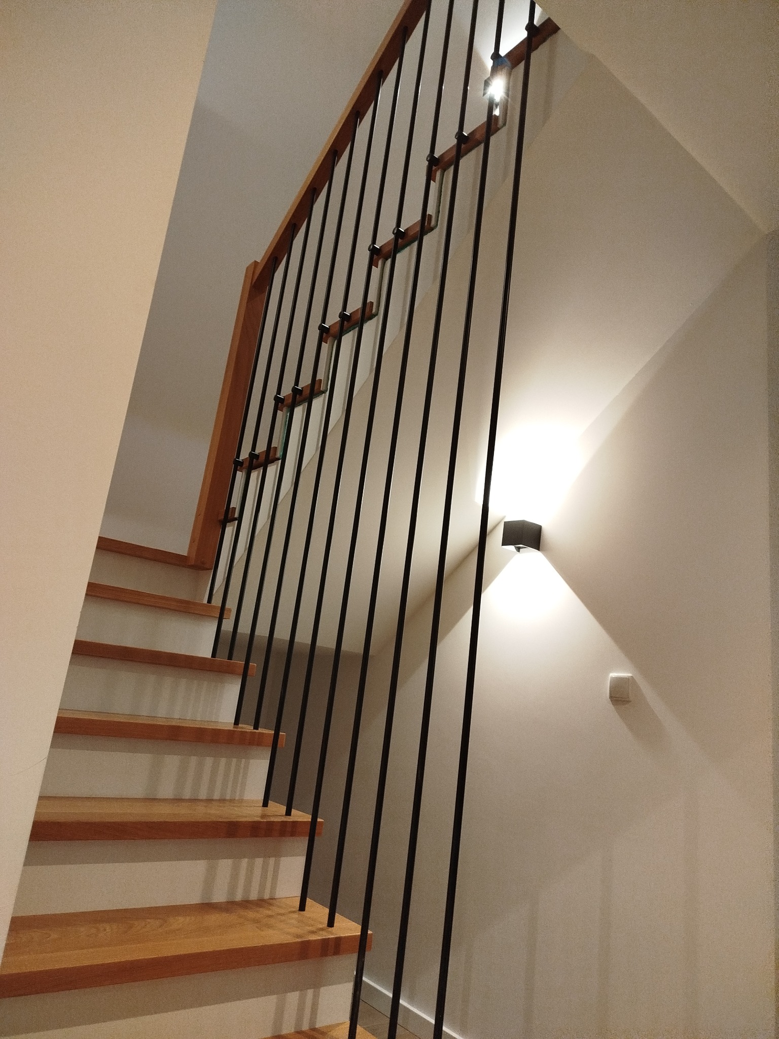 Nowoczesne schody z drewnianymi stopniami i czarną, minimalistyczną balustradą. Ściana oświetlona kinkietem, jasne wnętrze.