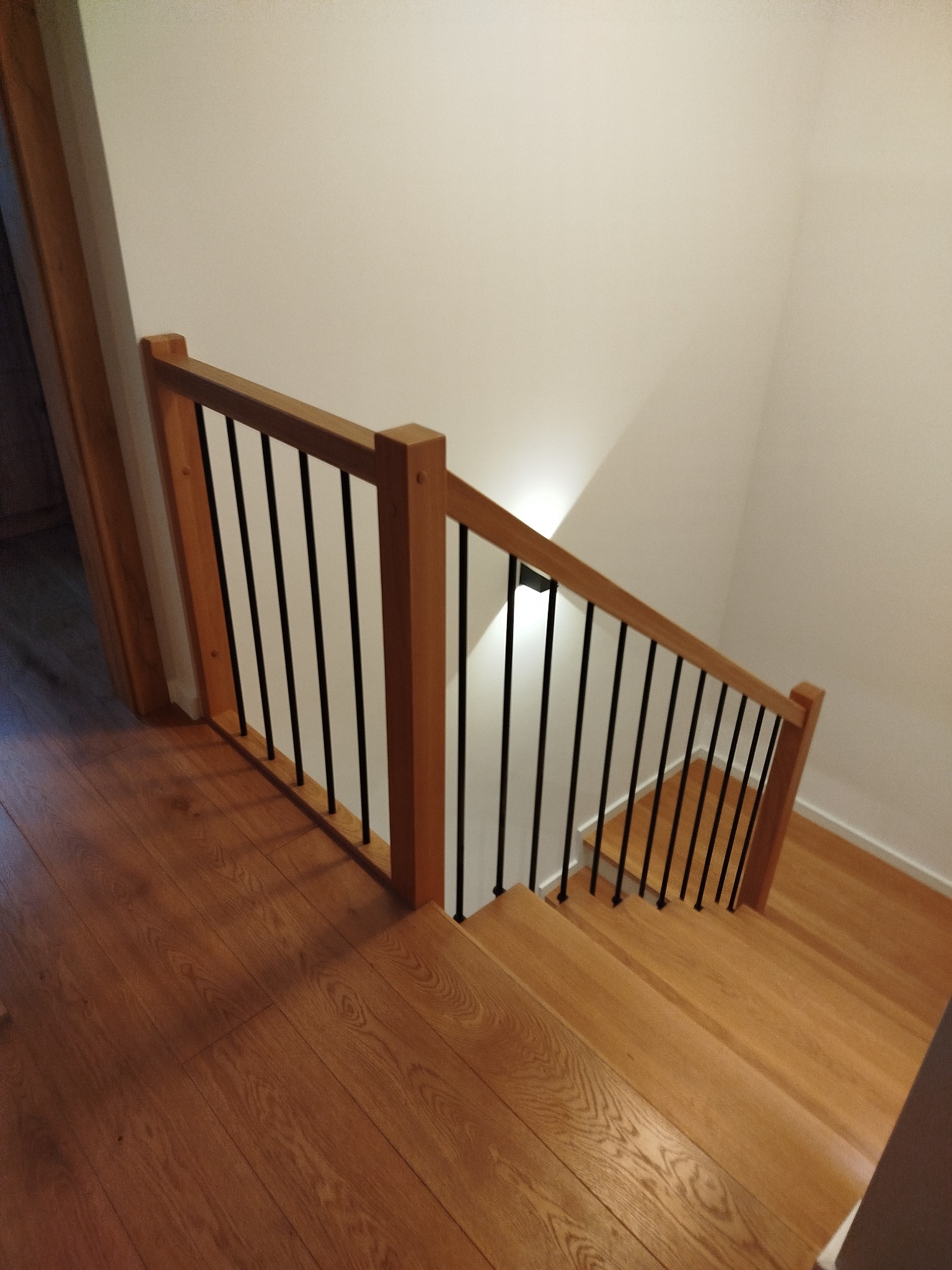 Jasne, drewniane schody z czarnymi, metalowymi balustradami, oświetlone punktowo, widziane z góry. Parkiet na podłodze. Minimalistyczny design.