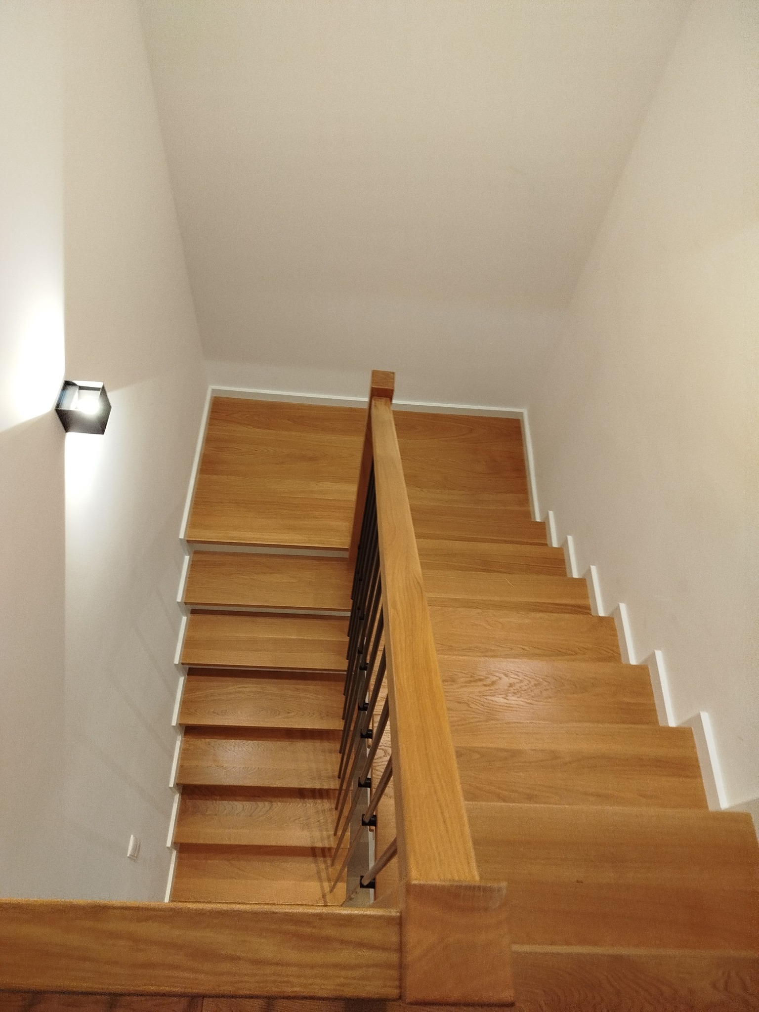 Jasne, dębowe stopnie z białymi podstopnicami i metalową balustradą, oświetlone nowoczesnym kinkietem, prowadzą w górę. Ujęcie z dołu, podkreślające solidność konstrukcji.