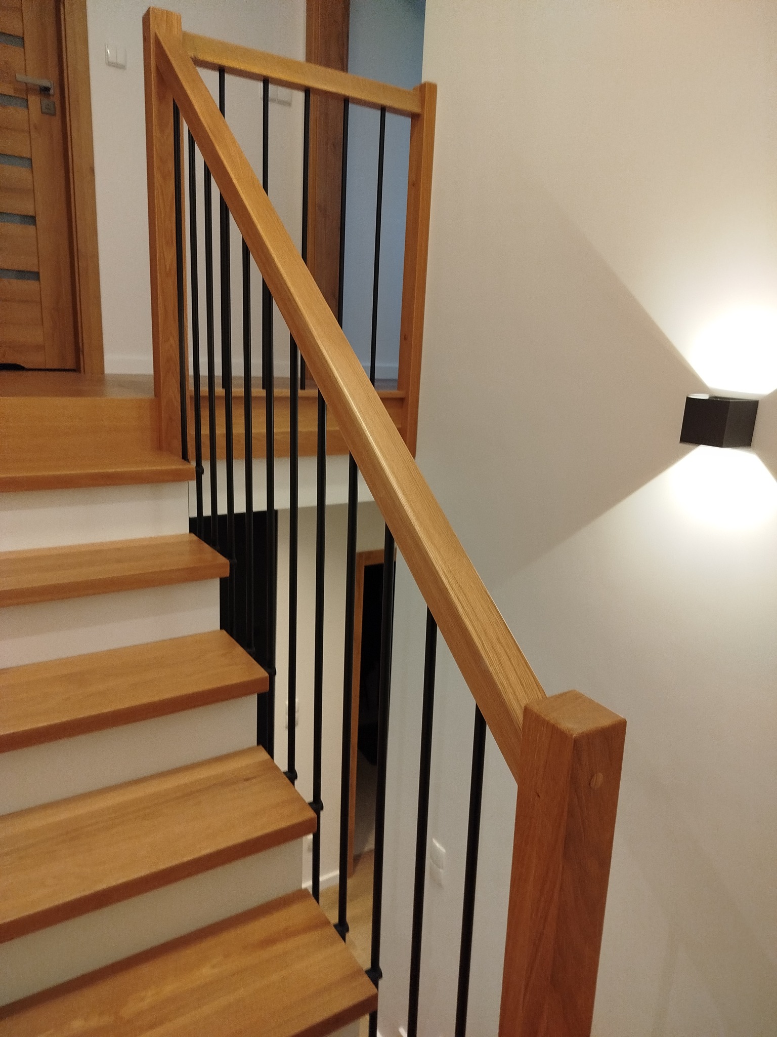 Jasne, drewniane stopnie z czarnymi, metalowymi balustradami i drewnianą poręczą, oświetlone nowoczesnym kinkietem na białej ścianie. Widok z dołu.