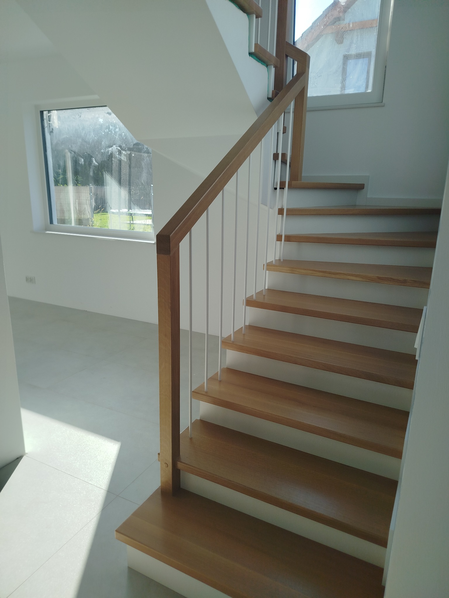 Jasne, nowoczesne schody z drewnianymi stopniami i białymi balustradami w minimalistycznym wnętrzu. Widoczne okno z refleksami światła. Ujęcie z dołu, podkreślające geometrię.
