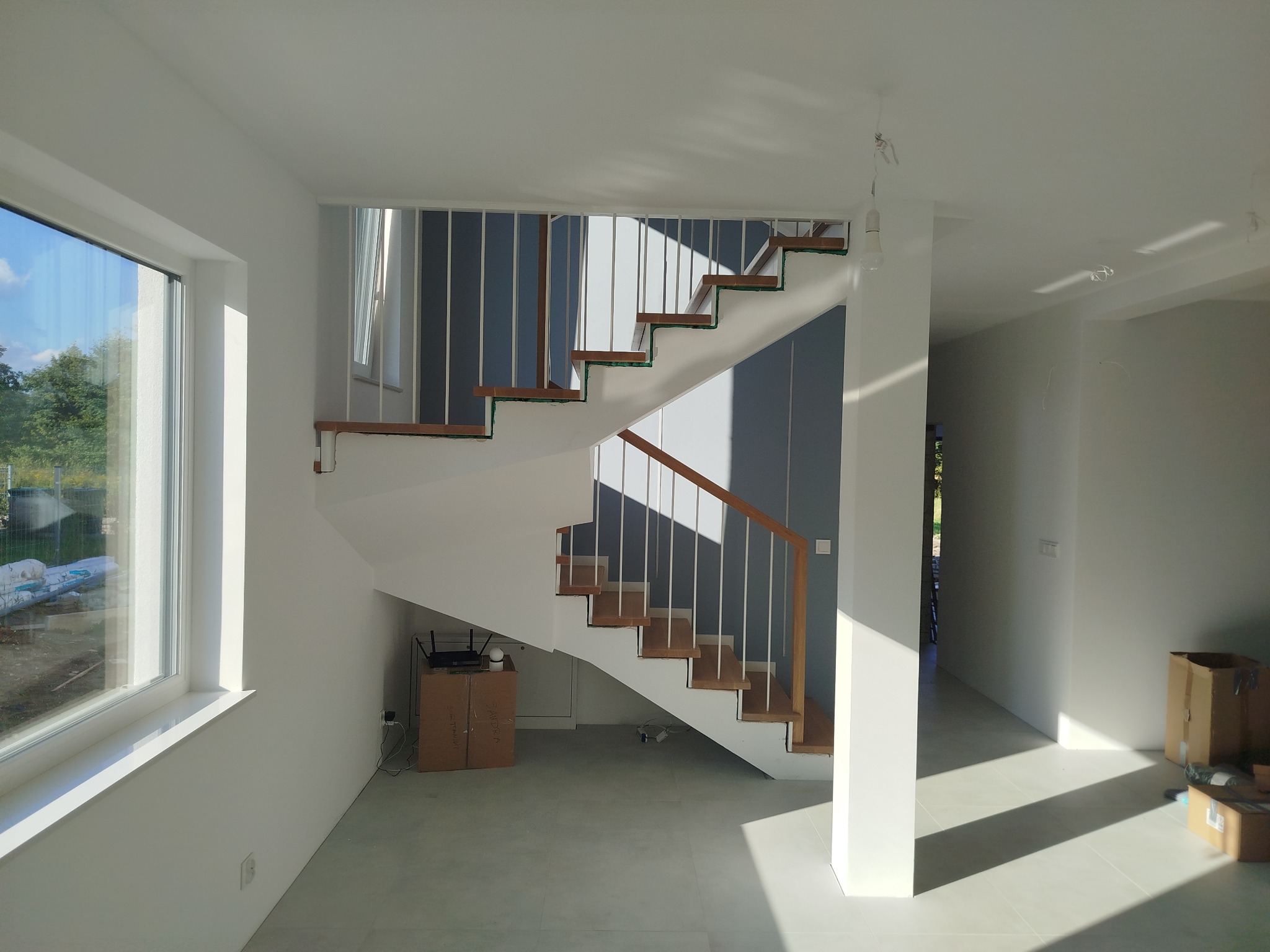 Nowoczesne schody z drewnianymi stopniami i białymi barierkami w jasnym wnętrzu. Widoczne duże okno i kartony, sugerujące świeżo zakończony montaż.