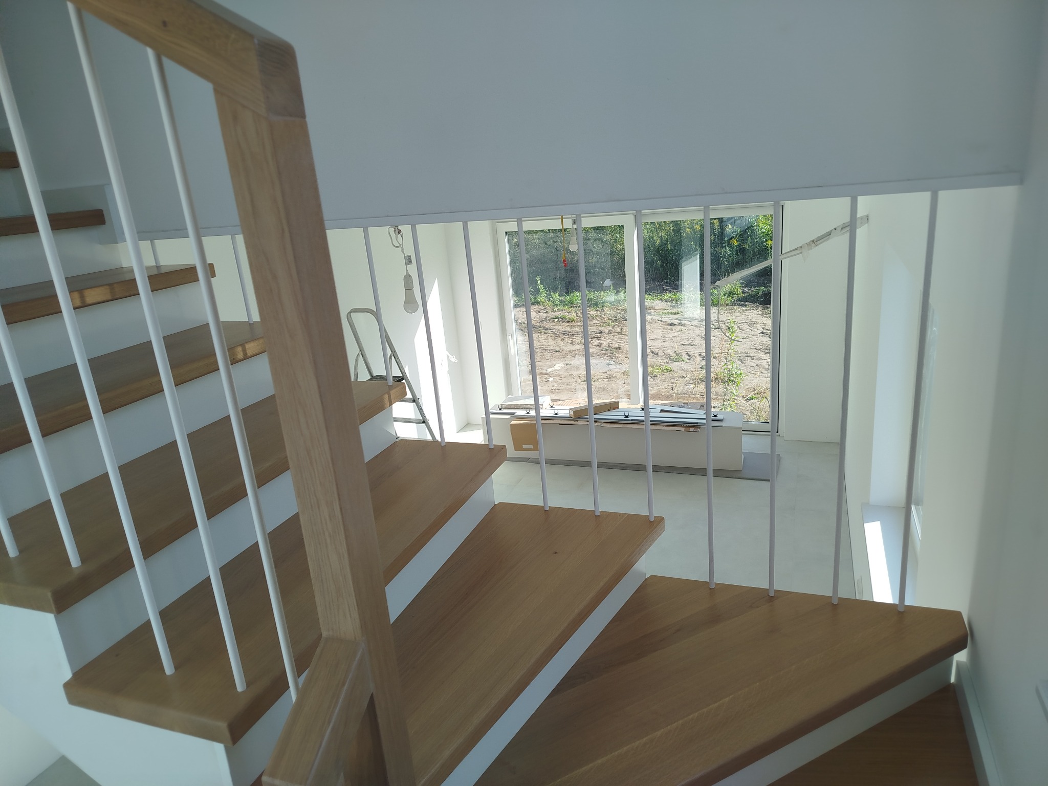 Jasne, nowoczesne schody z drewnianymi stopniami i białymi barierkami, widziane z góry. W tle okno z widokiem na zieleń.