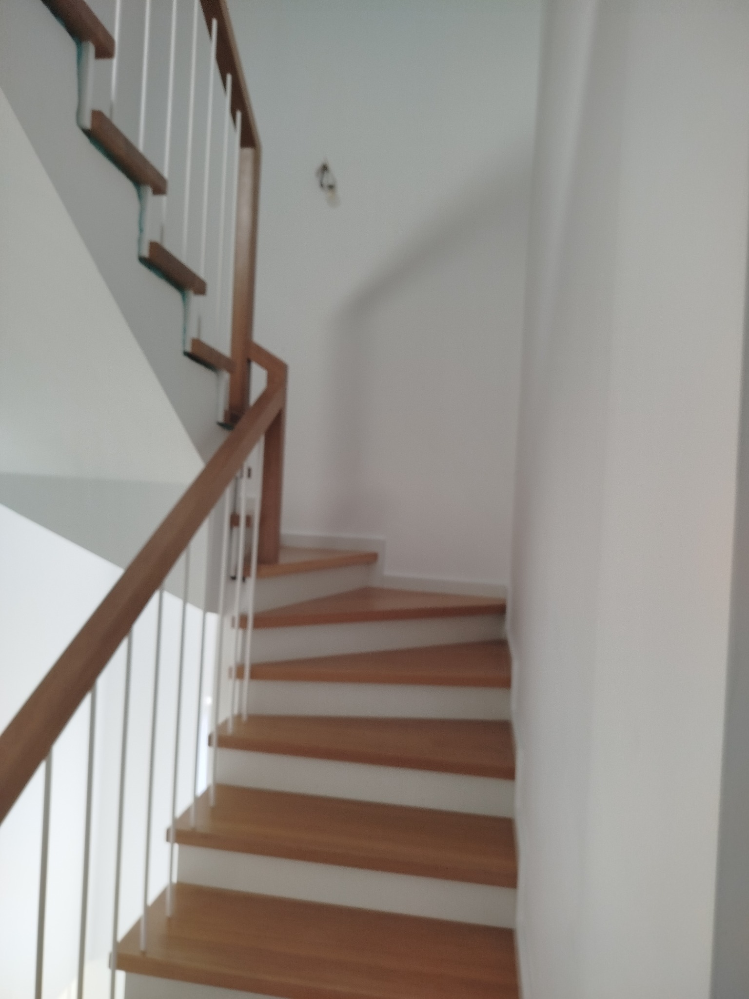 Jasne, drewniane stopnie z białymi podstopnicami i balustradą z drewnianą poręczą. Ujęcie od dołu, widok na zakręt schodów w nowoczesnym wnętrzu.