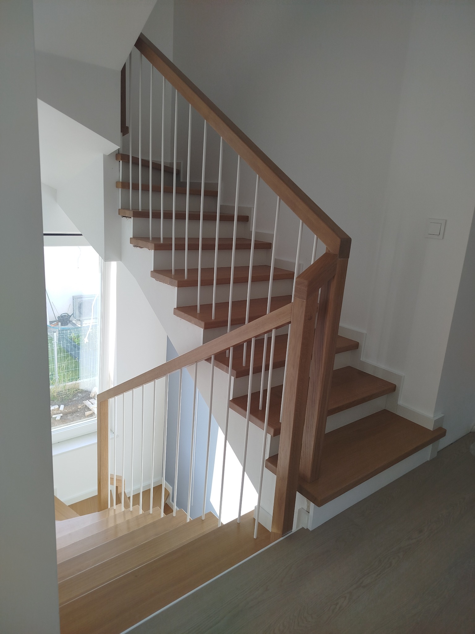 Jasne drewniane schody z białymi balustradami w nowoczesnym wnętrzu. Widok z góry, ukazujący detale wykończenia i naturalne światło wpadające przez okno.