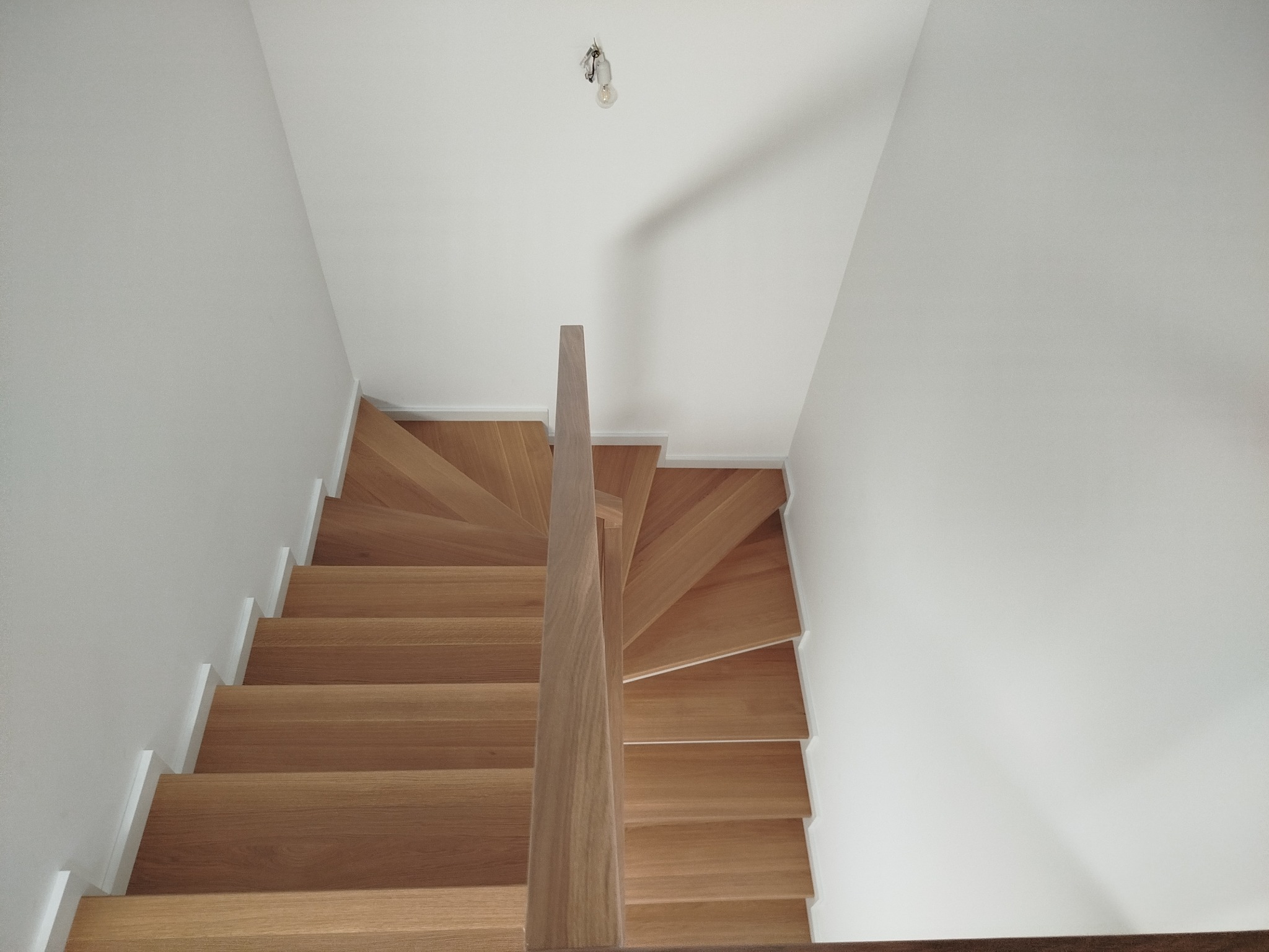 Jasne, drewniane schody z prostymi stopniami i minimalistyczną barierką, widziane z góry, w białym wnętrzu z żarówką na suficie.