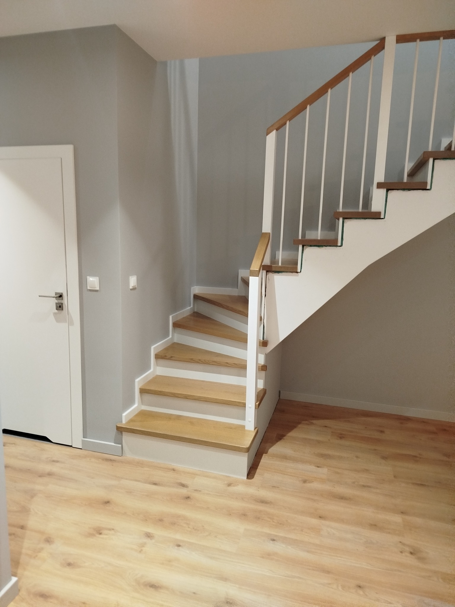 Jasne, nowoczesne schody z drewnianymi stopniami i białymi barierkami w szarej klatce schodowej. Widoczny fragment podłogi z paneli i białe drzwi po lewej.