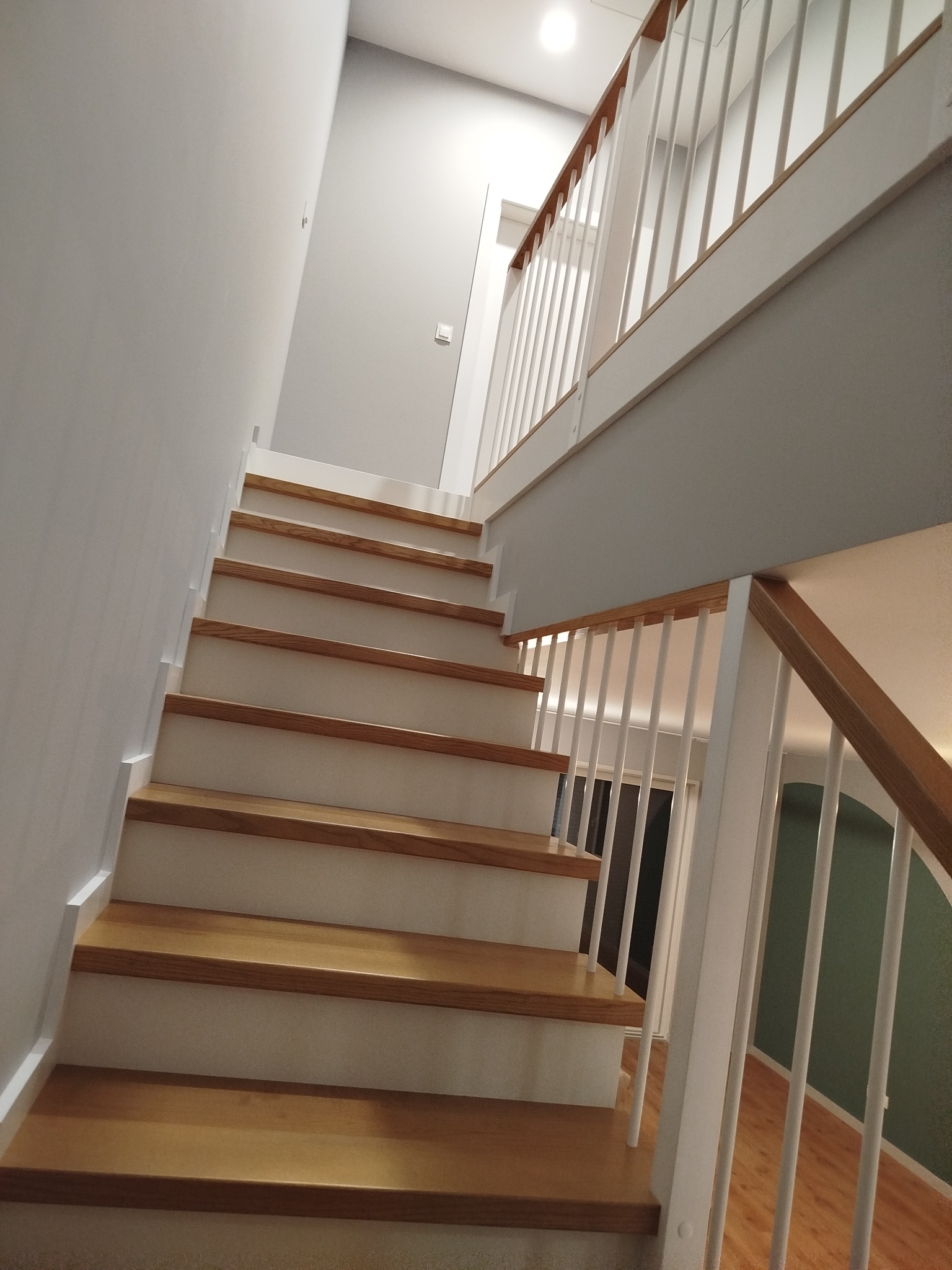 Jasne, drewniane stopnie schodów z białymi balustradami w nowoczesnym wnętrzu. Widok z dołu, podkreślający pionowe linie balustrad i ciepło drewna.