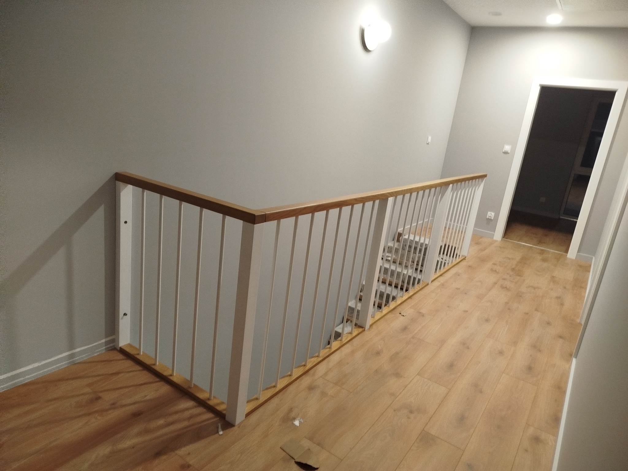 Drewniane schody z białą balustradą i drewnianym poręczą, widok z góry na drewnianą podłogę i szare ściany. Nowoczesny design, oświetlenie punktowe.