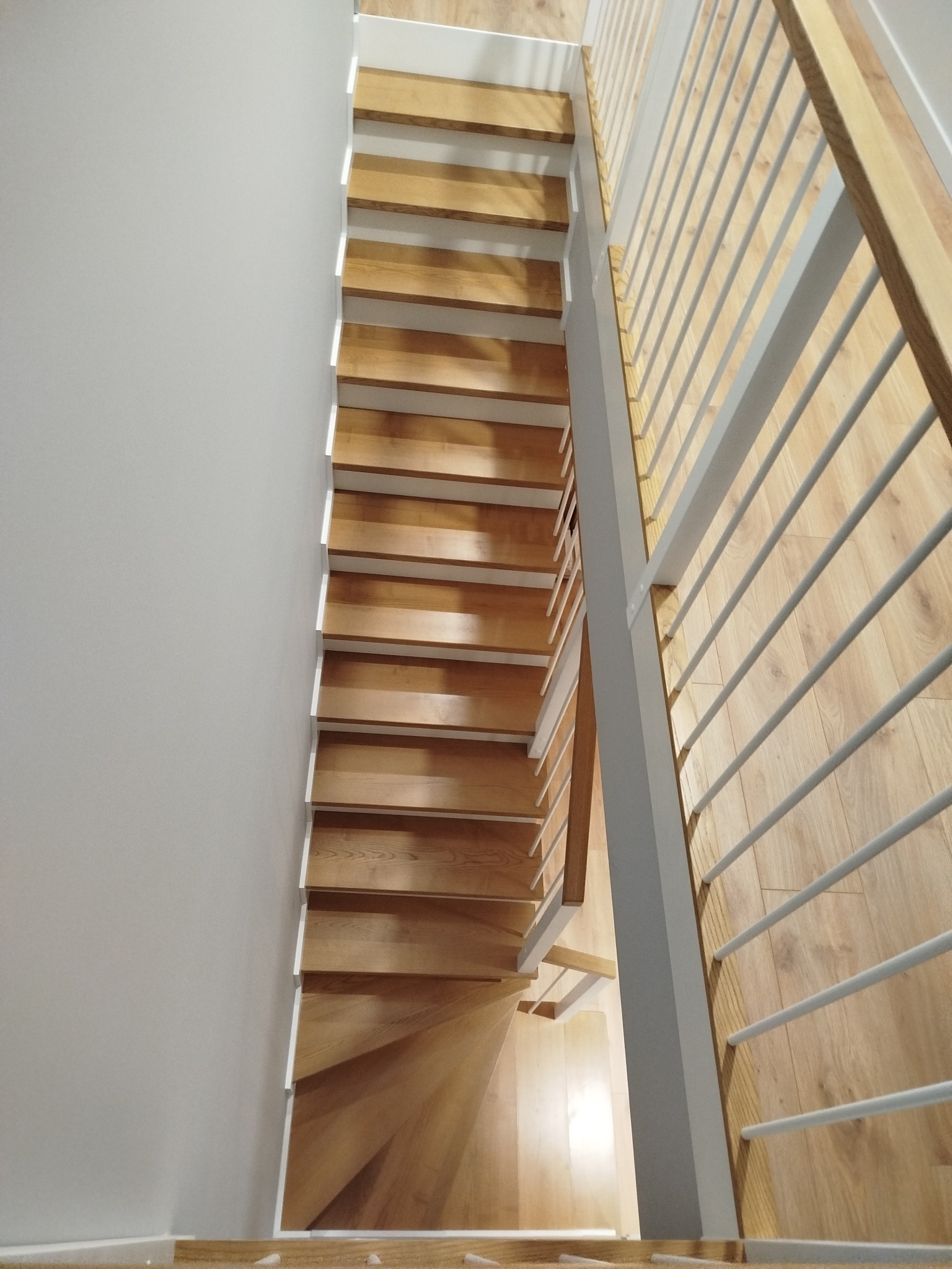 Jasne, drewniane stopnie schodów z białymi podstopnicami, widziane z góry, z metalową balustradą po prawej stronie. Minimalistyczny design, naturalne światło.