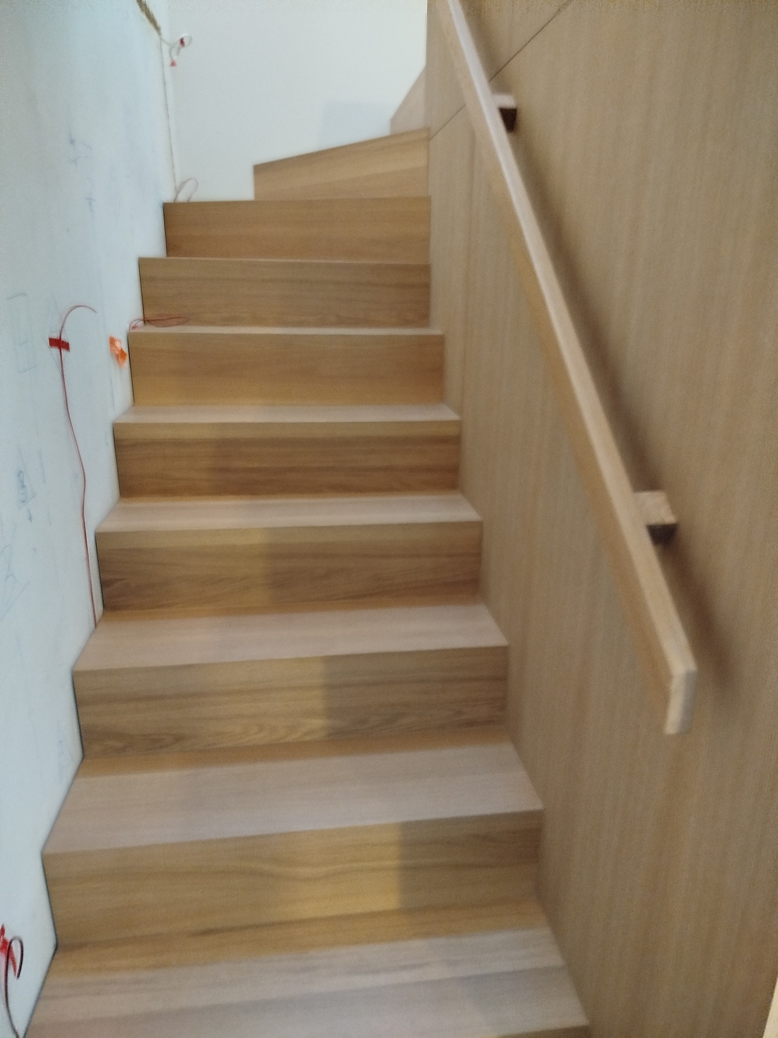 Jasne, drewniane stopnie schodów z prostą balustradą, widoczne przewody na ścianie, sugerujące etap wykończeniowy. Minimalistyczny design.