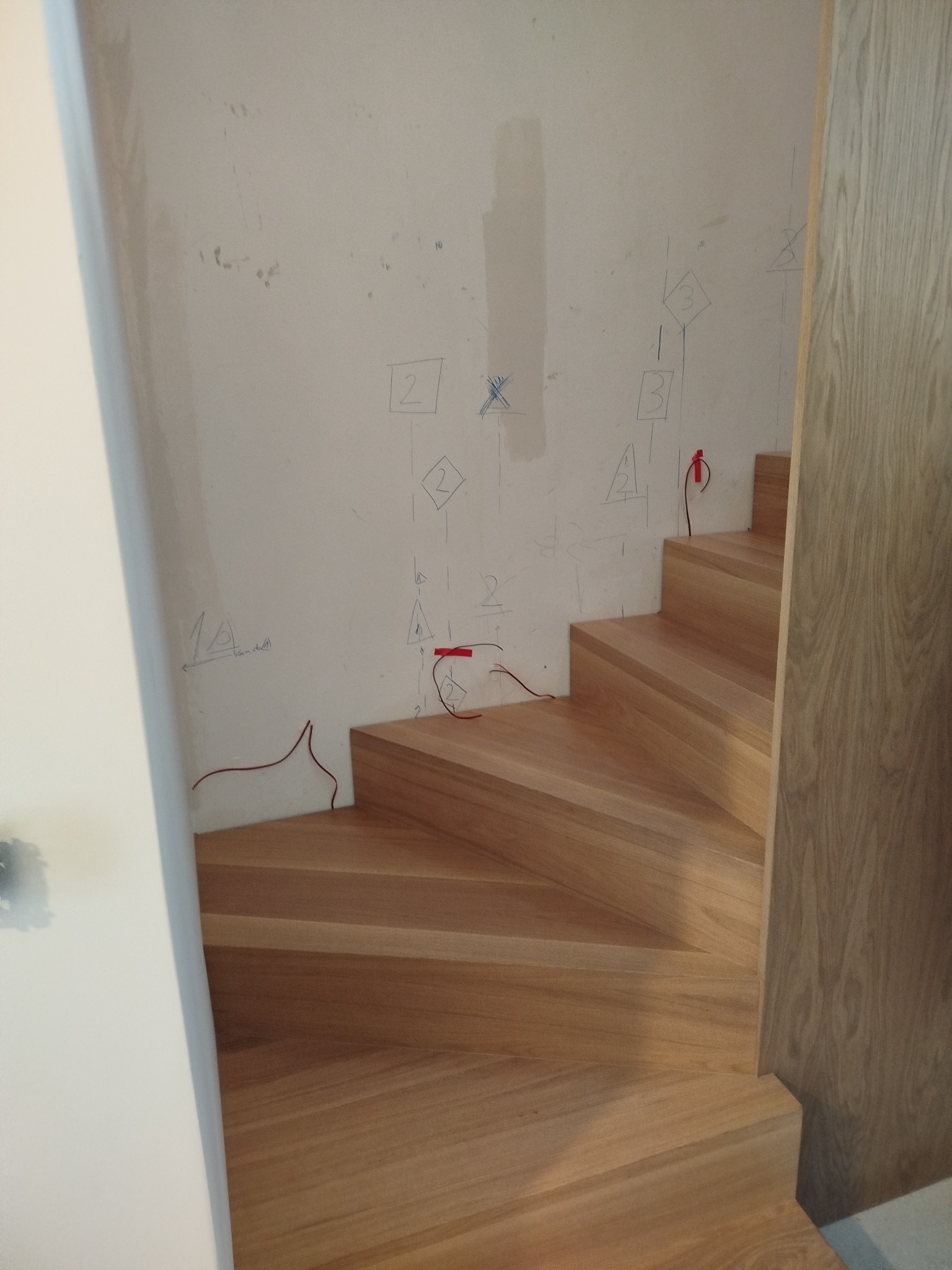 Jasne, drewniane schody spiralne w trakcie montażu, widoczne oznaczenia na ścianie i wystające przewody elektryczne. Ujęcie z dołu, podkreślające kształt.