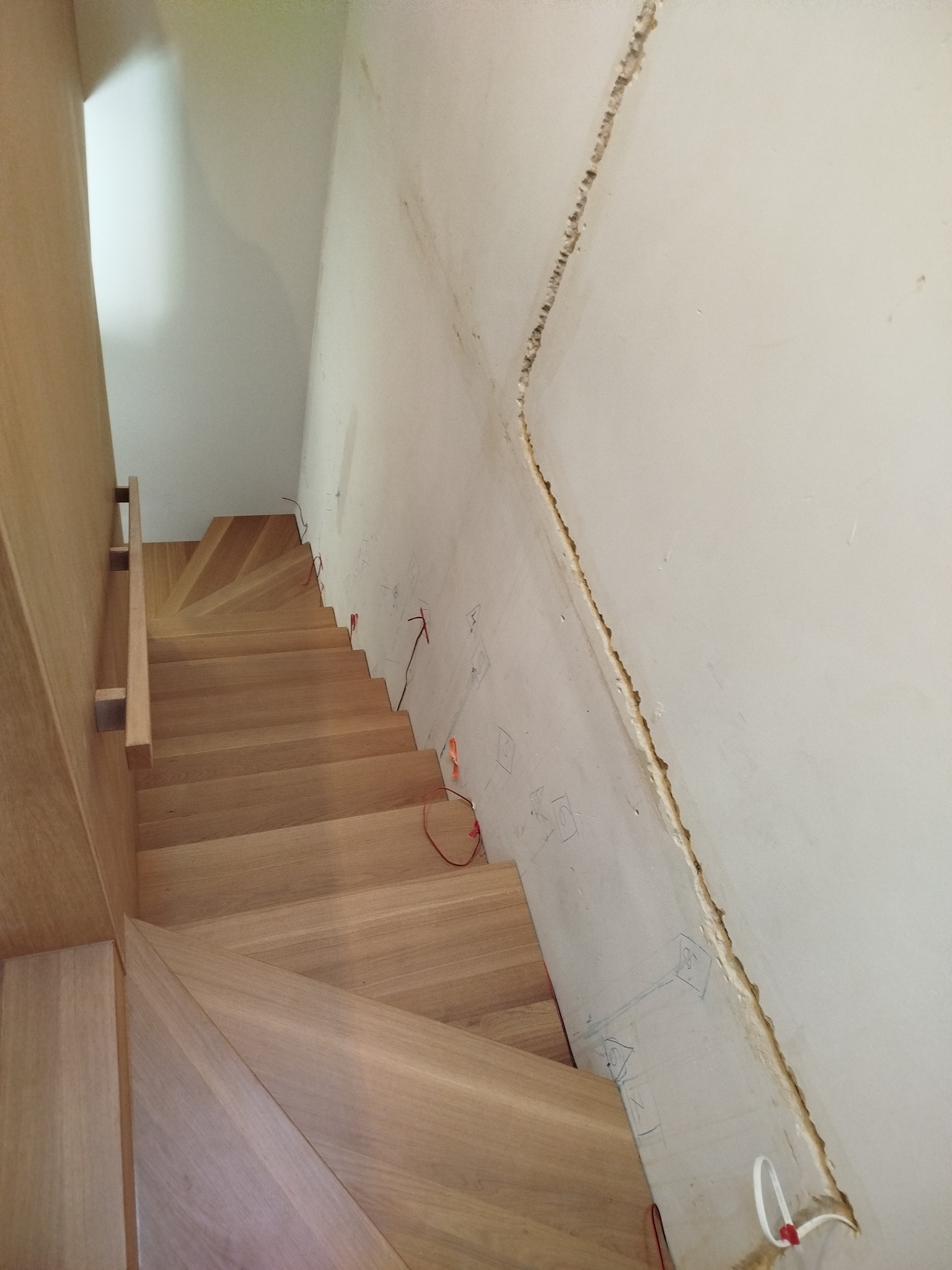 Montaż jasnych, drewnianych schodów z balustradą w surowym wnętrzu. Widoczne przygotowania do wykończenia ściany, zarysowane punkty elektryczne i kable.