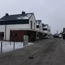 Rząd nowo wybudowanych, dwukondygnacyjnych domów szeregowych z ciemnymi dachami i drewnianymi panelami na elewacji, widziany z brukowanej drogi w pochmurny dzień z niewielką ilością śniegu.