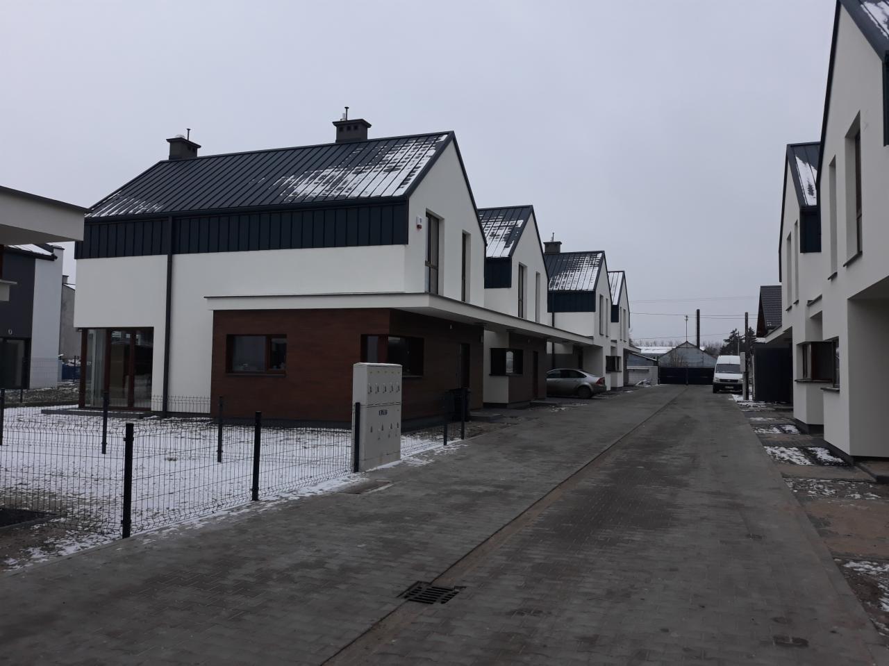 Rząd nowo wybudowanych, dwukondygnacyjnych domów szeregowych z ciemnymi dachami i drewnianymi panelami na elewacji, widziany z brukowanej drogi w pochmurny dzień z niewielką ilością śniegu.