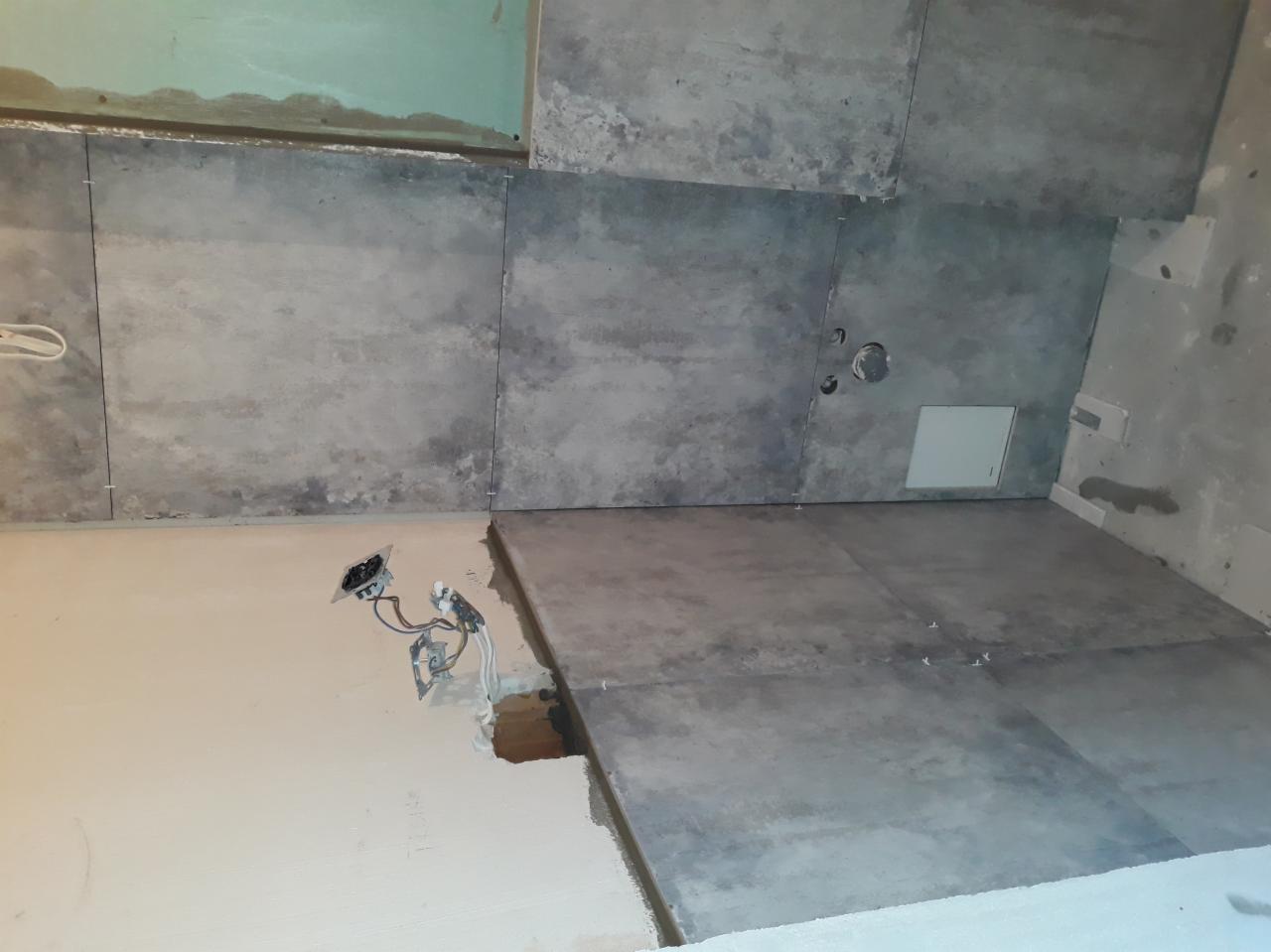 Układanie szarych płytek imitujących beton na ścianie i podłodze w trakcie prac wykończeniowych, widoczne przewody elektryczne i niedokończone ściany.