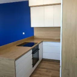 Nowoczesna kuchnia z białymi szafkami i drewnianym blatem, niebieska ściana w tle, płyta indukcyjna i piekarnik w zabudowie.