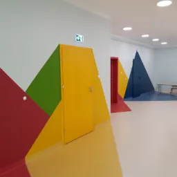 Kolorowy korytarz z geometrycznymi wzorami na ścianach, żółte i czerwone drzwi, jasna podłoga, widok na dalszą część korytarza z ławkami.