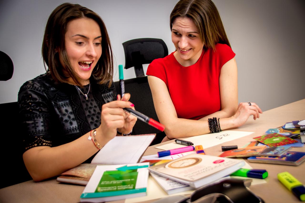 Dwie kobiety przy biurku, jedna z otwartymi ustami trzyma markery, druga uśmiecha się, na biurku notatniki, książki i kolorowe kartki.