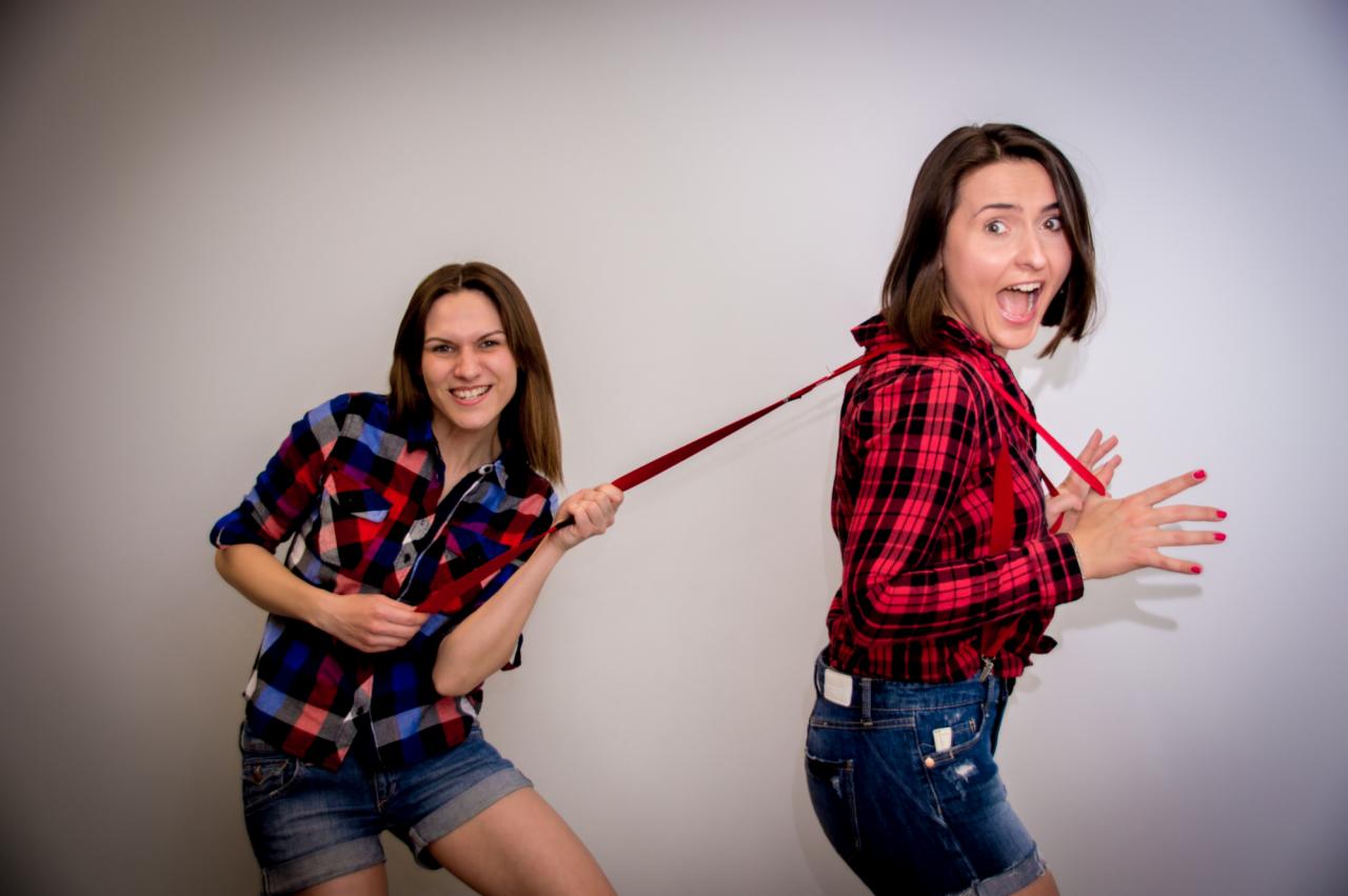 Dwie kobiety w koszulach w kratę i jeansowych szortach, jedna ciągnie drugą za czerwoną tasiemkę z zaskoczoną miną na jasnoszarym tle.