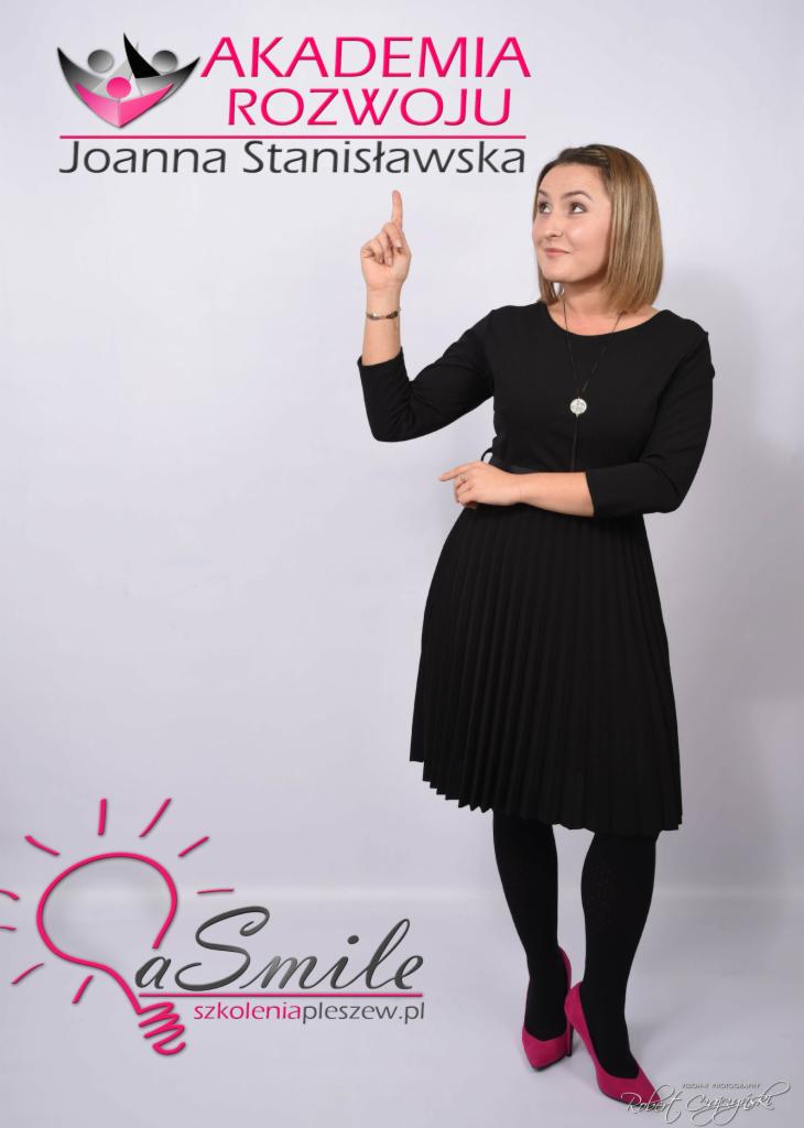 Kobieta w czarnej sukience i różowych szpilkach, wskazująca palcem w górę na tle logo firmy szkoleniowej.