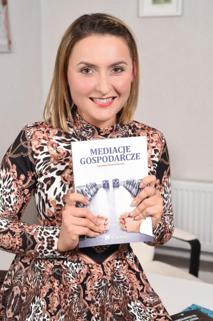 Uśmiechnięta kobieta w wzorzystej sukience prezentuje książkę 'Mediacje Gospodarcze' z ilustracją ścieżki na okładce.