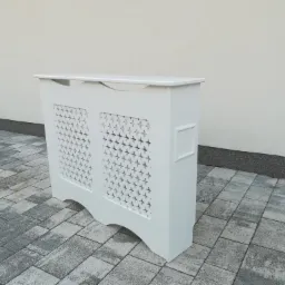 Biała, dekoracyjna obudowa na grzejnik z ażurowym wzorem krzyżyków, ustawiona na szarej kostce brukowej przed jasną ścianą budynku.