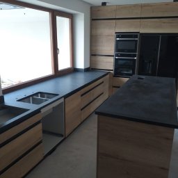 K-Stolar - Nowoczesna kuchnia z drewnianymi szafkami i czarnym blatem, wyspa kuchenna z blatem w tym samym stylu. Widoczne okno nad zlewem i zabudowane sprzęty AGD.