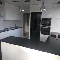 K-Stolar - Nowoczesna kuchnia z białymi szafkami i czarnym blatem, oświetlona srebrnymi lampami wiszącymi. Widoczny sprzęt AGD w zabudowie i marmurowa podłoga.