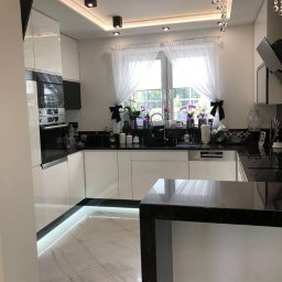 K-Stolar - Nowoczesna kuchnia z białymi szafkami, czarnym blatem i wyspą. Oświetlenie LED pod szafkami i marmurowe płytki podłogowe. Okno z firankami i dekoracjami.