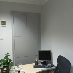 K-Stolar - Szara szafa wnękowa z czterema drzwiami, wkomponowana w ścianę biura. Na biurku monitor, klawiatura, roślina doniczkowa i butelka wody. Minimalistyczny design.