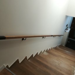 K-Stolar - Drewniane schody z zamontowaną, jasną poręczą z ciemnymi zakończeniami, przymocowaną do białej ściany. Widok z góry na dół, w tle drzwi wejściowe.