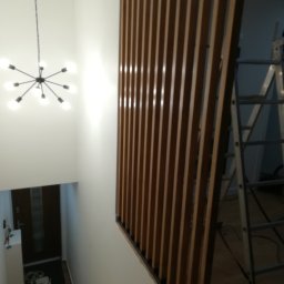 K-Stolar - Nowoczesna balustrada z drewnianych listew w Mielcu, oddzielająca schody od przestrzeni. Widoczny fragment schodów i lampa sufitowa. W tle drabina.