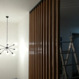 K-Stolar - Drewniana ścianka działowa z pionowych listew w nowoczesnym wnętrzu. W tle widoczna drabina, sugerująca prace wykończeniowe. Minimalistyczny design z lampą w stylu industrialnym.