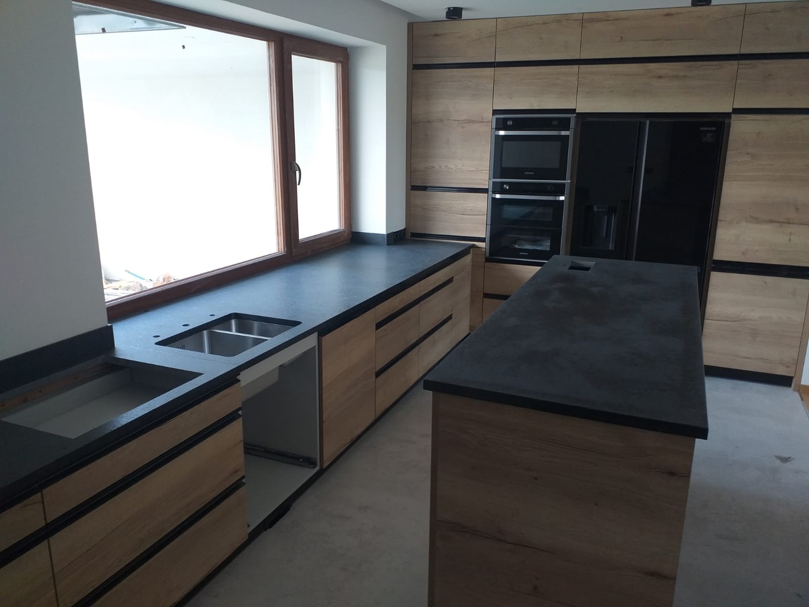 Nowoczesna kuchnia z drewnianymi szafkami i czarnym blatem, wyspa kuchenna z blatem w tym samym stylu. Widoczne okno nad zlewem i zabudowane sprzęty AGD.