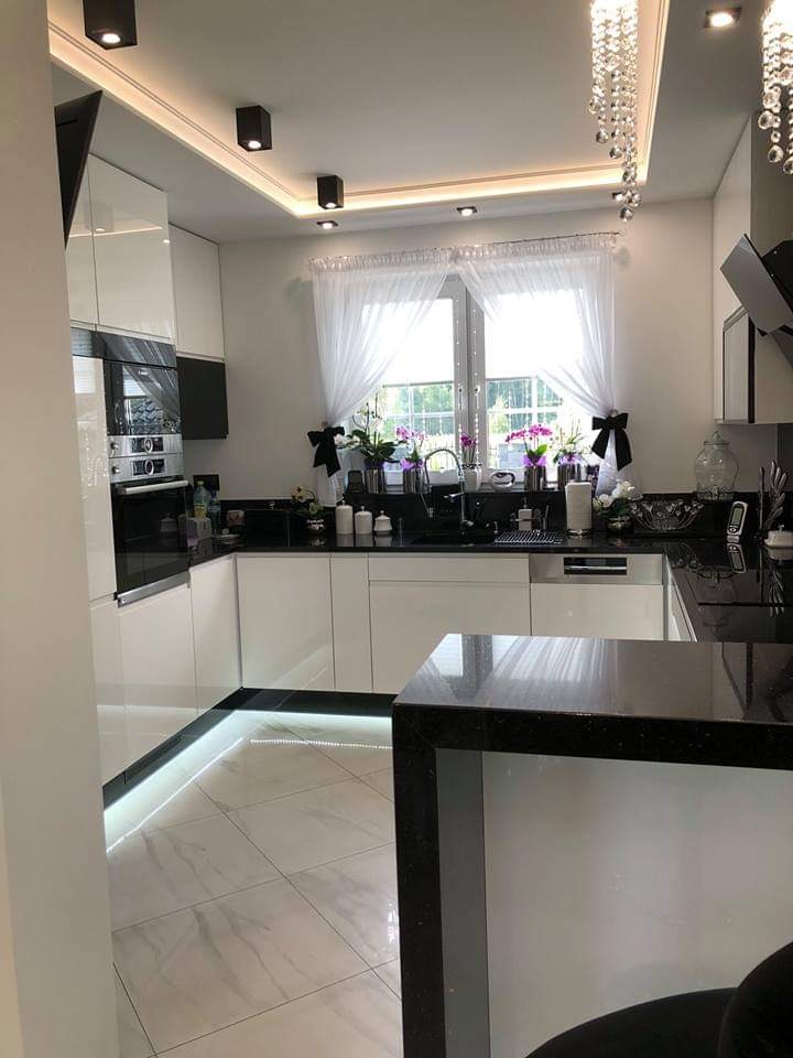 Nowoczesna kuchnia z białymi szafkami, czarnym blatem i wyspą. Oświetlenie LED pod szafkami i marmurowe płytki podłogowe. Okno z firankami i dekoracjami.