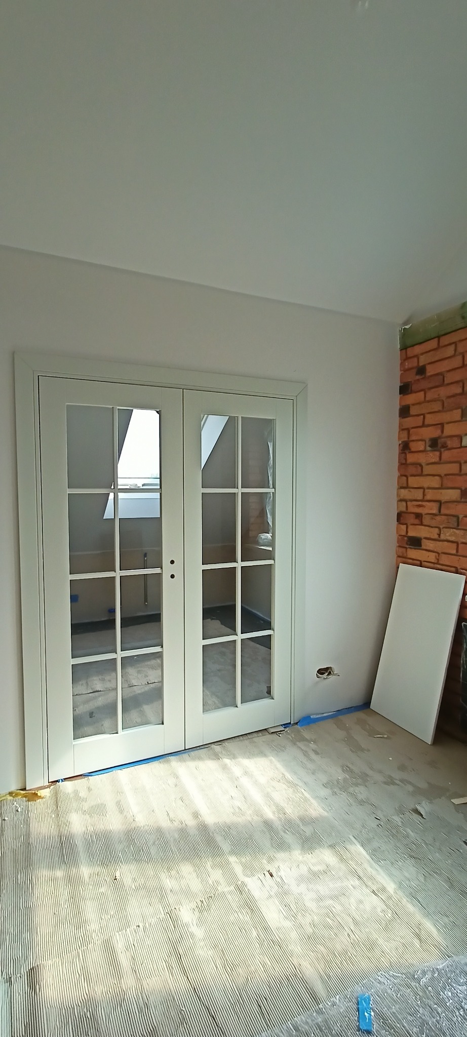 Białe, przeszklone drzwi dwuskrzydłowe z podziałami, zamontowane w jasnym pomieszczeniu z ceglaną ścianą. Widoczne przygotowania do położenia podłogi.