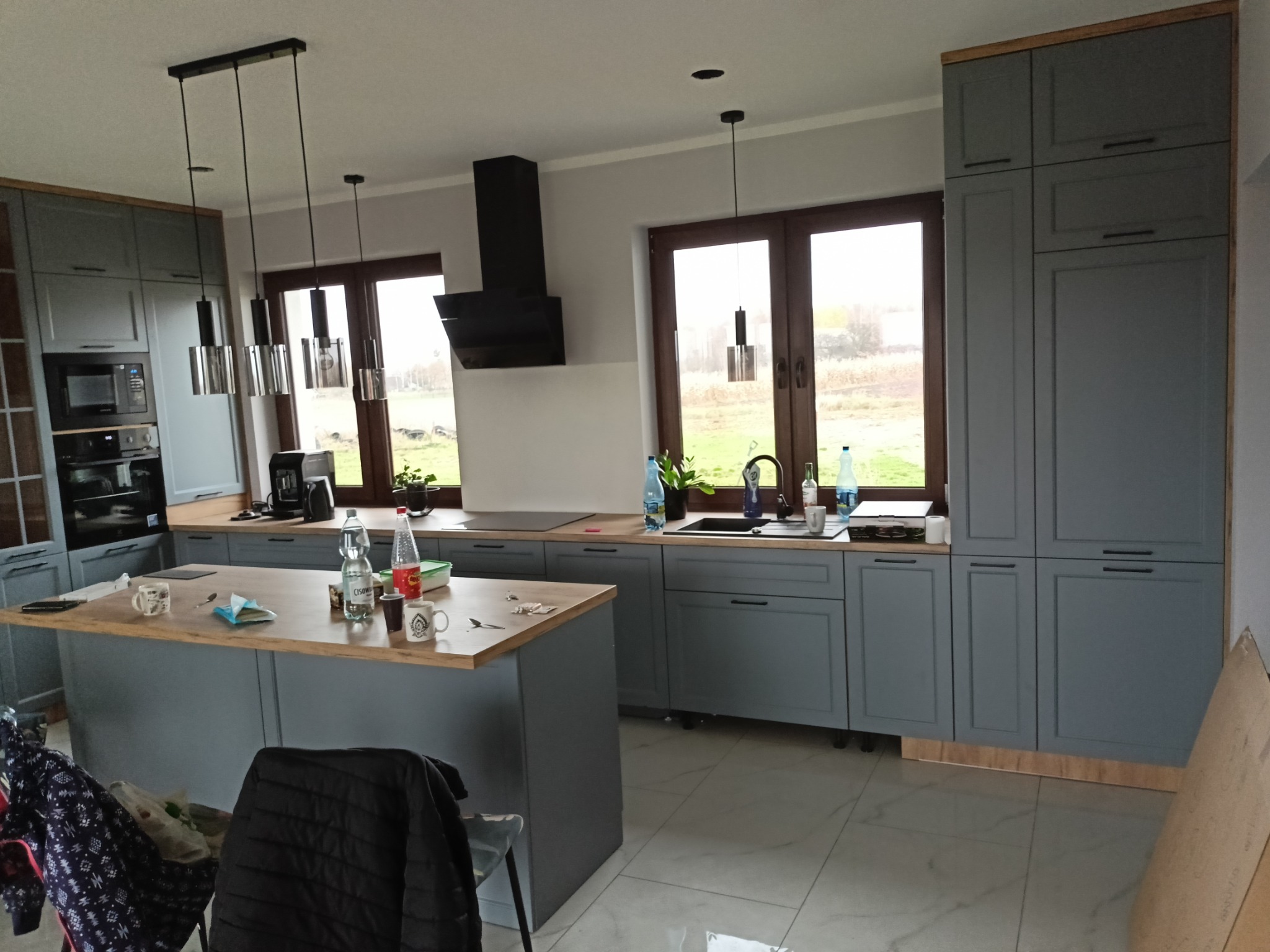 Szara kuchnia na wymiar z drewnianym blatem i wyspą, oświetlona wiszącymi lampami. Widok na okno i ogród w tle. Elementy wyposażenia na blacie wyspy.