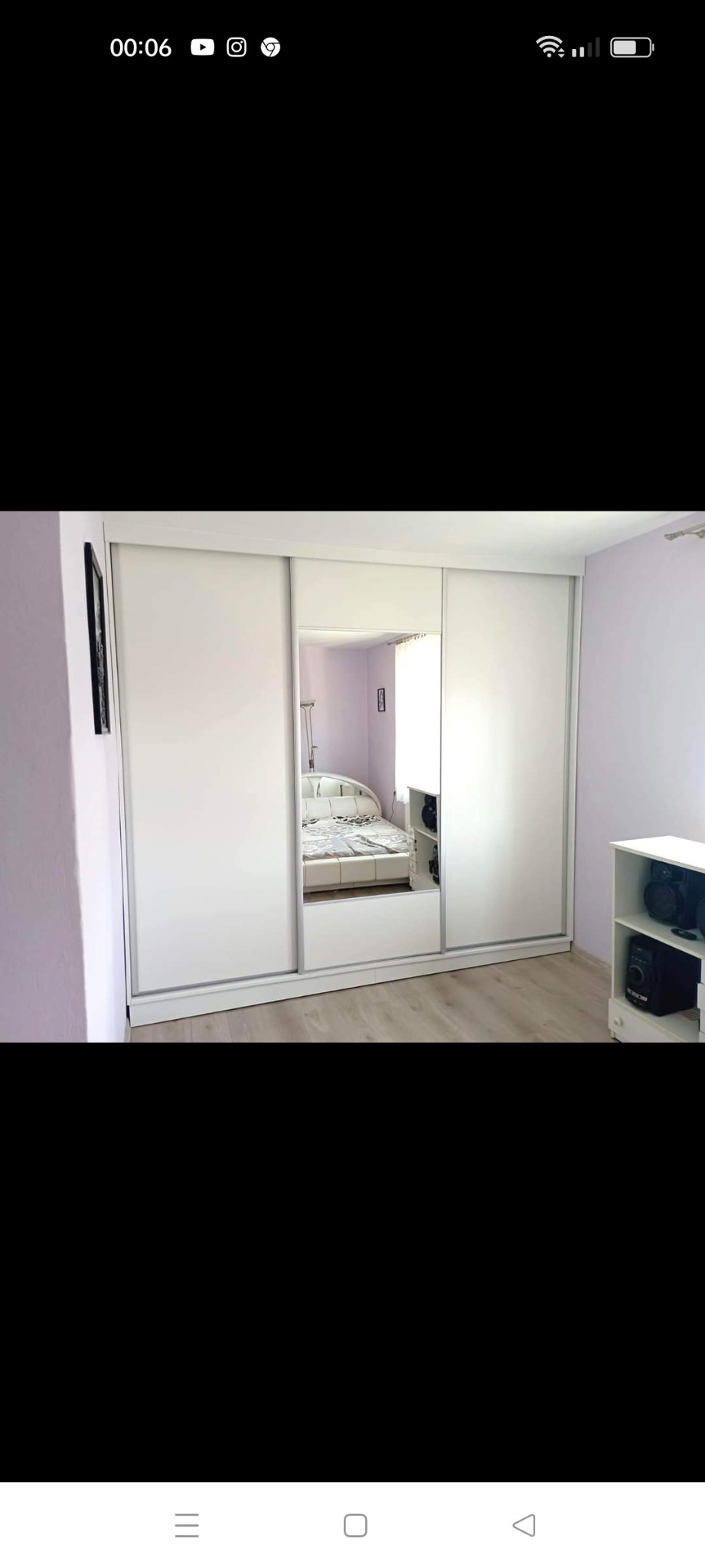 Biała szafa wnękowa z przesuwnymi drzwiami i lustrem, odbijająca wnętrze sypialni. Widoczna podłoga z paneli i fragment ściany w odcieniach fioletu.