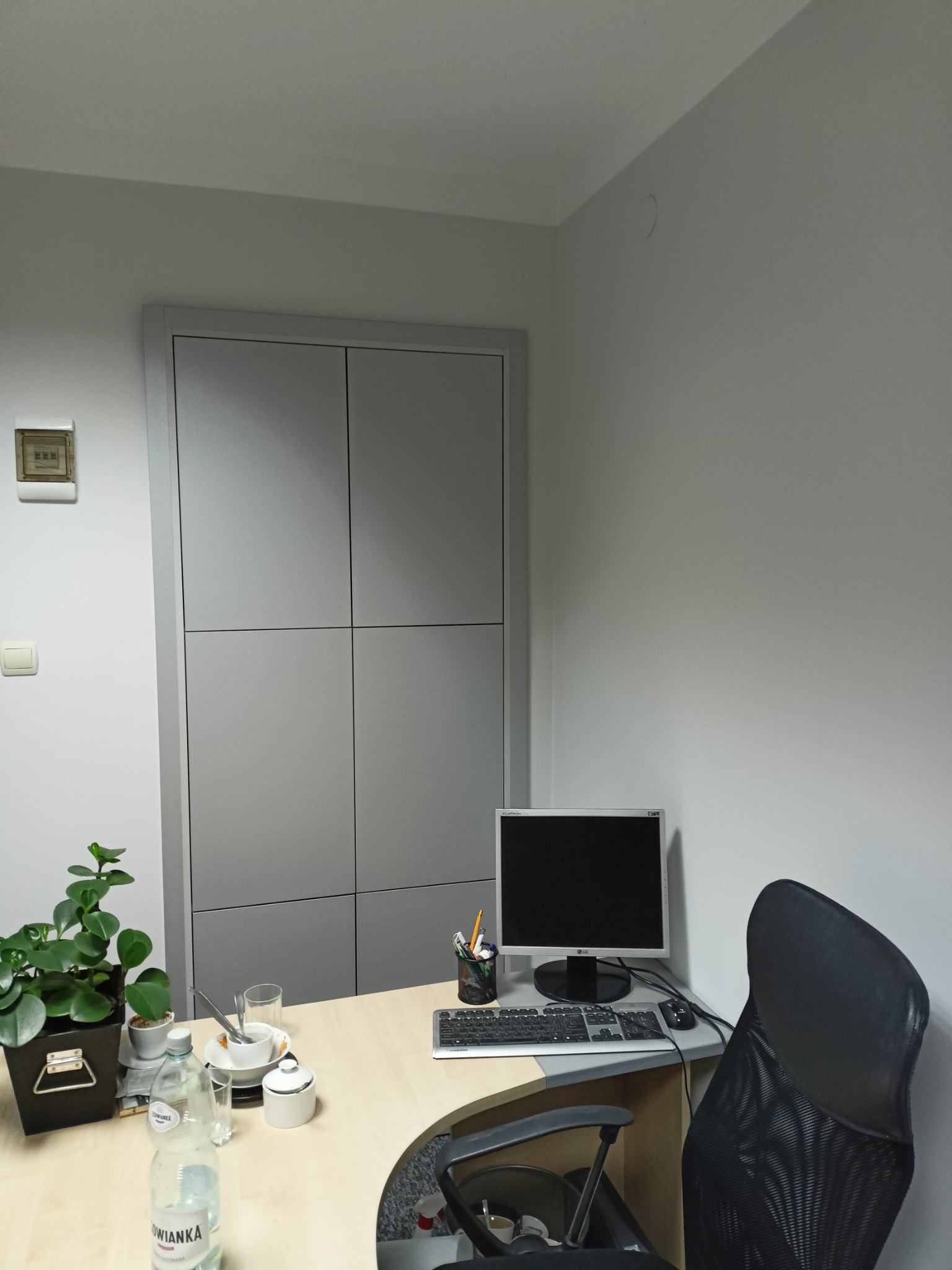 Szara szafa wnękowa z czterema drzwiami, wkomponowana w ścianę biura. Na biurku monitor, klawiatura, roślina doniczkowa i butelka wody. Minimalistyczny design.