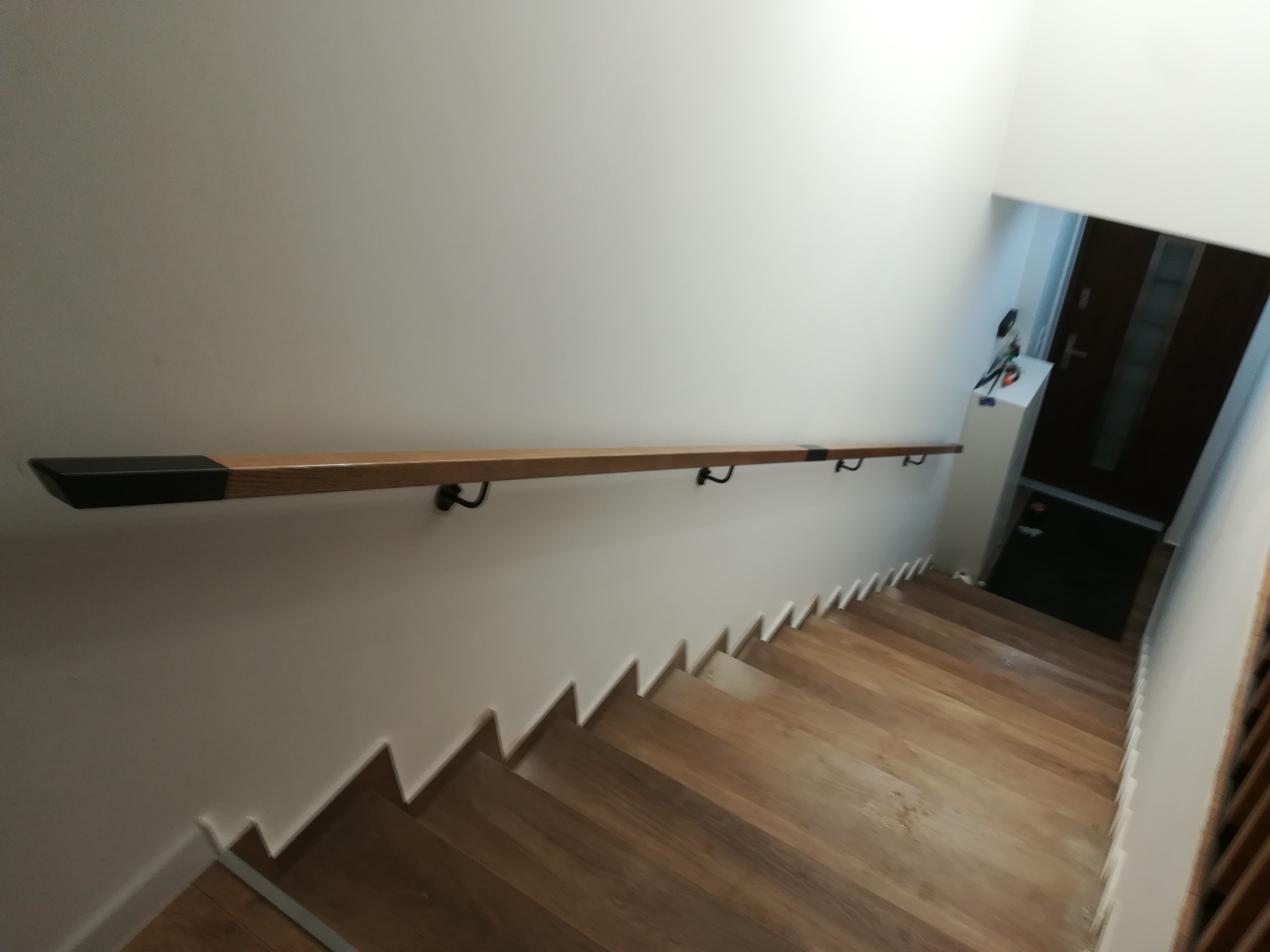 Drewniane schody z zamontowaną, jasną poręczą z ciemnymi zakończeniami, przymocowaną do białej ściany. Widok z góry na dół, w tle drzwi wejściowe.