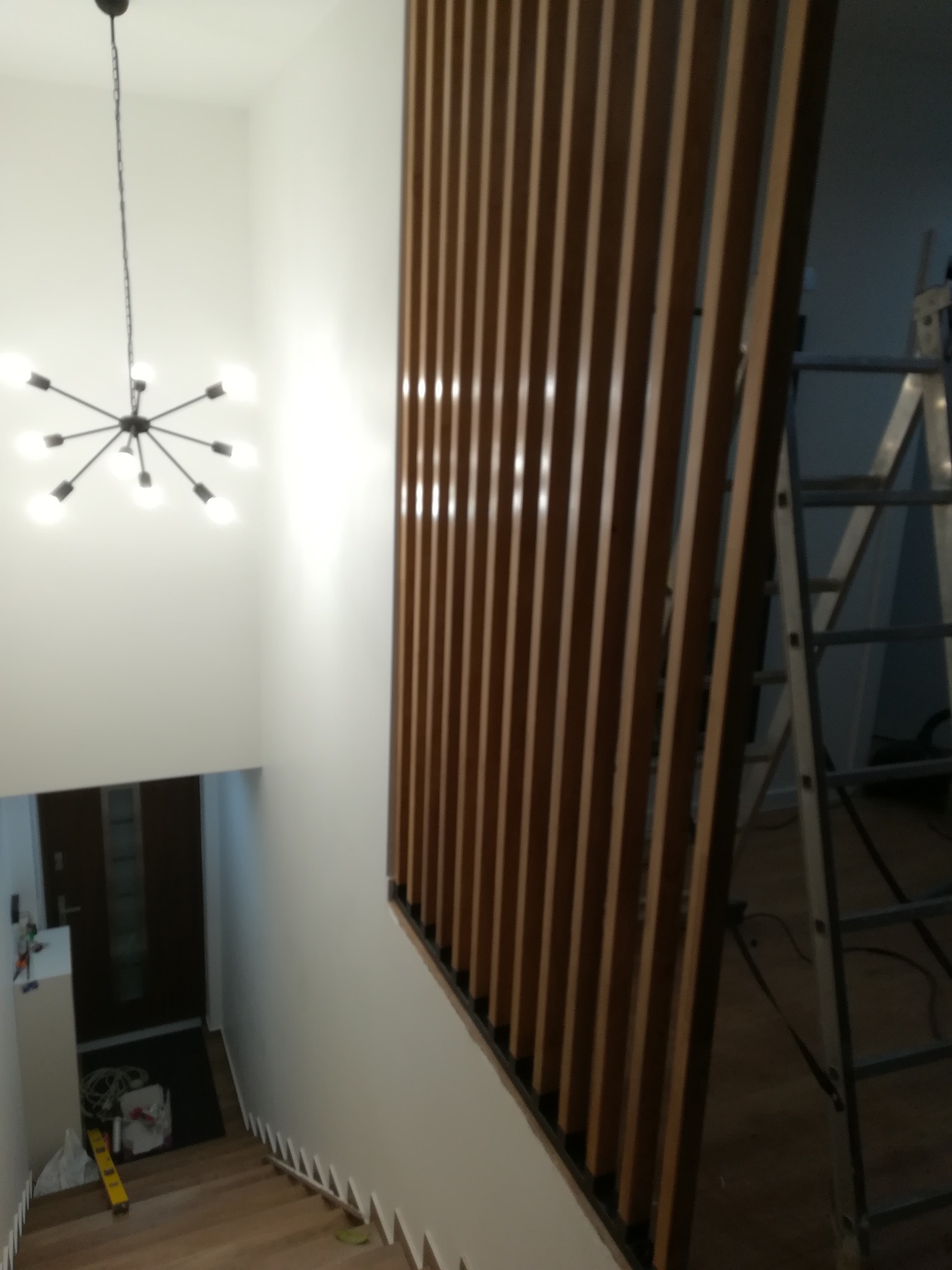 Nowoczesna balustrada z drewnianych listew w Mielcu, oddzielająca schody od przestrzeni. Widoczny fragment schodów i lampa sufitowa. W tle drabina.