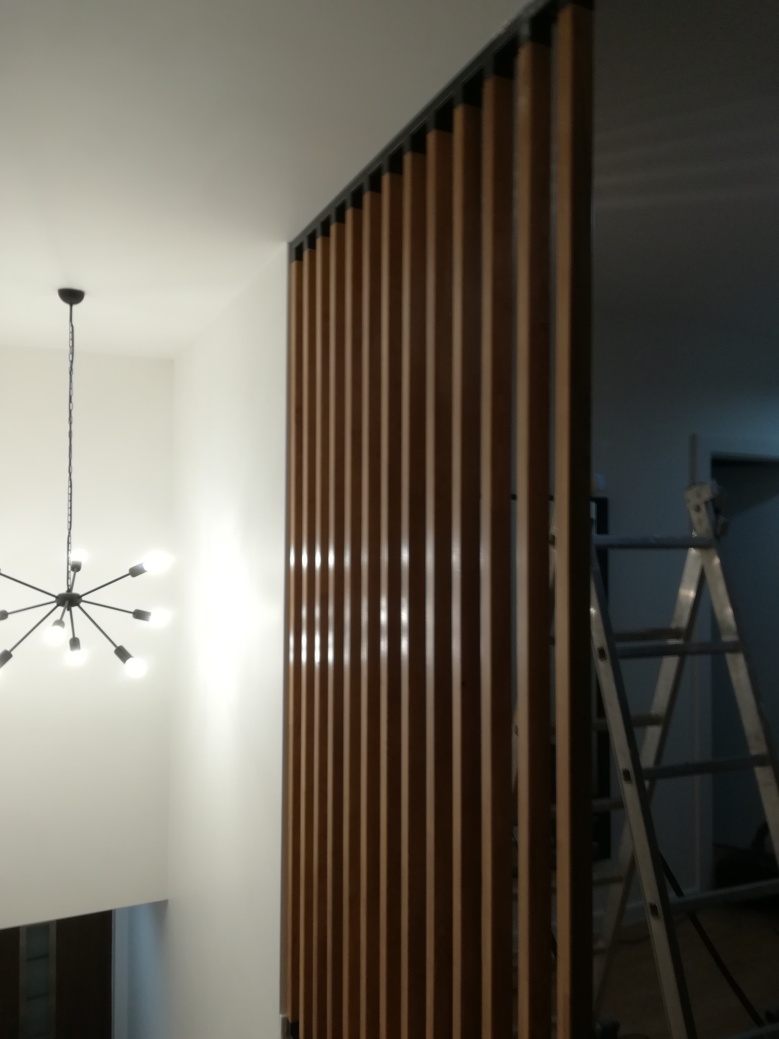 Drewniana ścianka działowa z pionowych listew w nowoczesnym wnętrzu. W tle widoczna drabina, sugerująca prace wykończeniowe. Minimalistyczny design z lampą w stylu industrialnym.