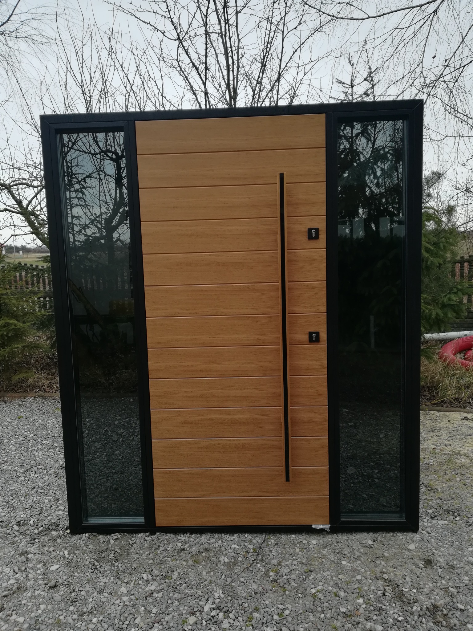 Nowoczesne drzwi zewnętrzne z drewnopodobnym panelem i czarną ramą, flankowane szybami. Minimalistyczny design, widoczne detale zamków i długa, pionowa klamka.
