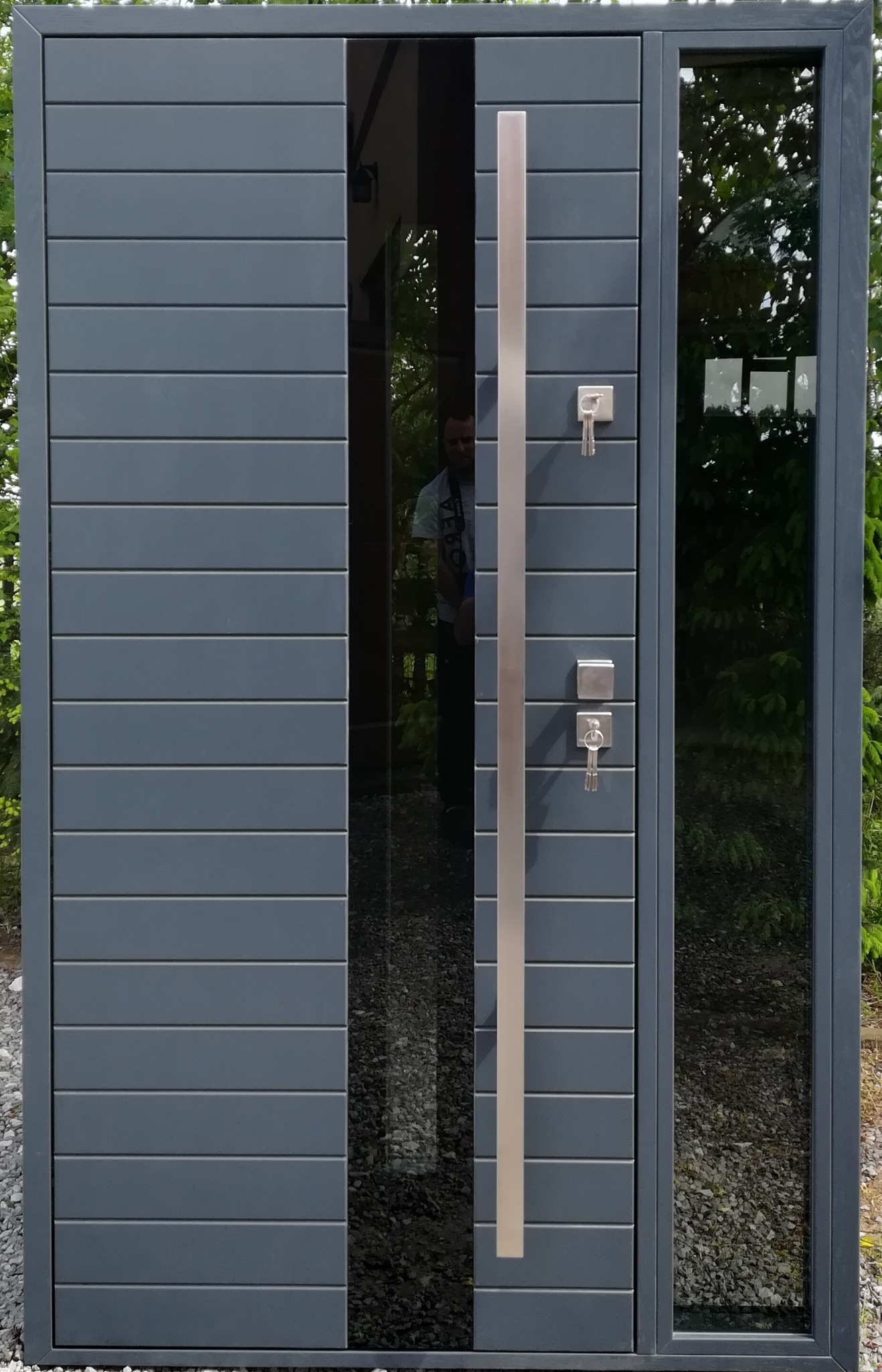 Nowoczesne, grafitowe drzwi zewnętrzne z poziomymi panelami, długim, srebrnym uchwytem i szybami po bokach, odbijające otoczenie. Klucze w zamku.
