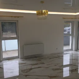 Puste, jasne wnętrze z marmurową podłogą, złotym żyrandolem i karniszami, grzejnikiem oraz dużymi oknami z widokiem na zewnątrz.