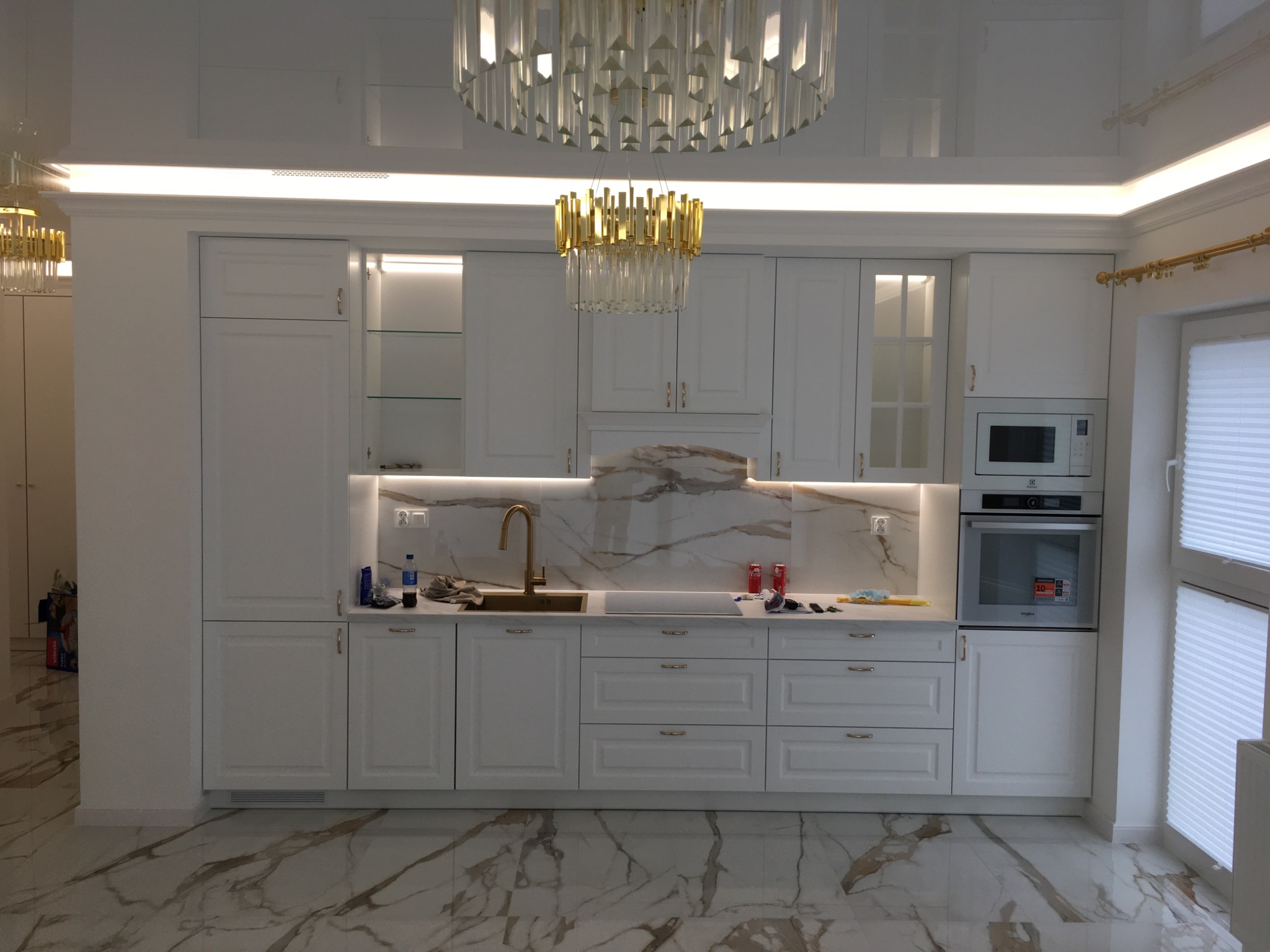 Elegancka, biała kuchnia z marmurowym blatem i backsplashiem, oświetleniem LED pod szafkami, oraz dwoma wiszącymi, kryształowymi żyrandolami, z widoczną armaturą w kolorze złotym i marmurową podłogą.
